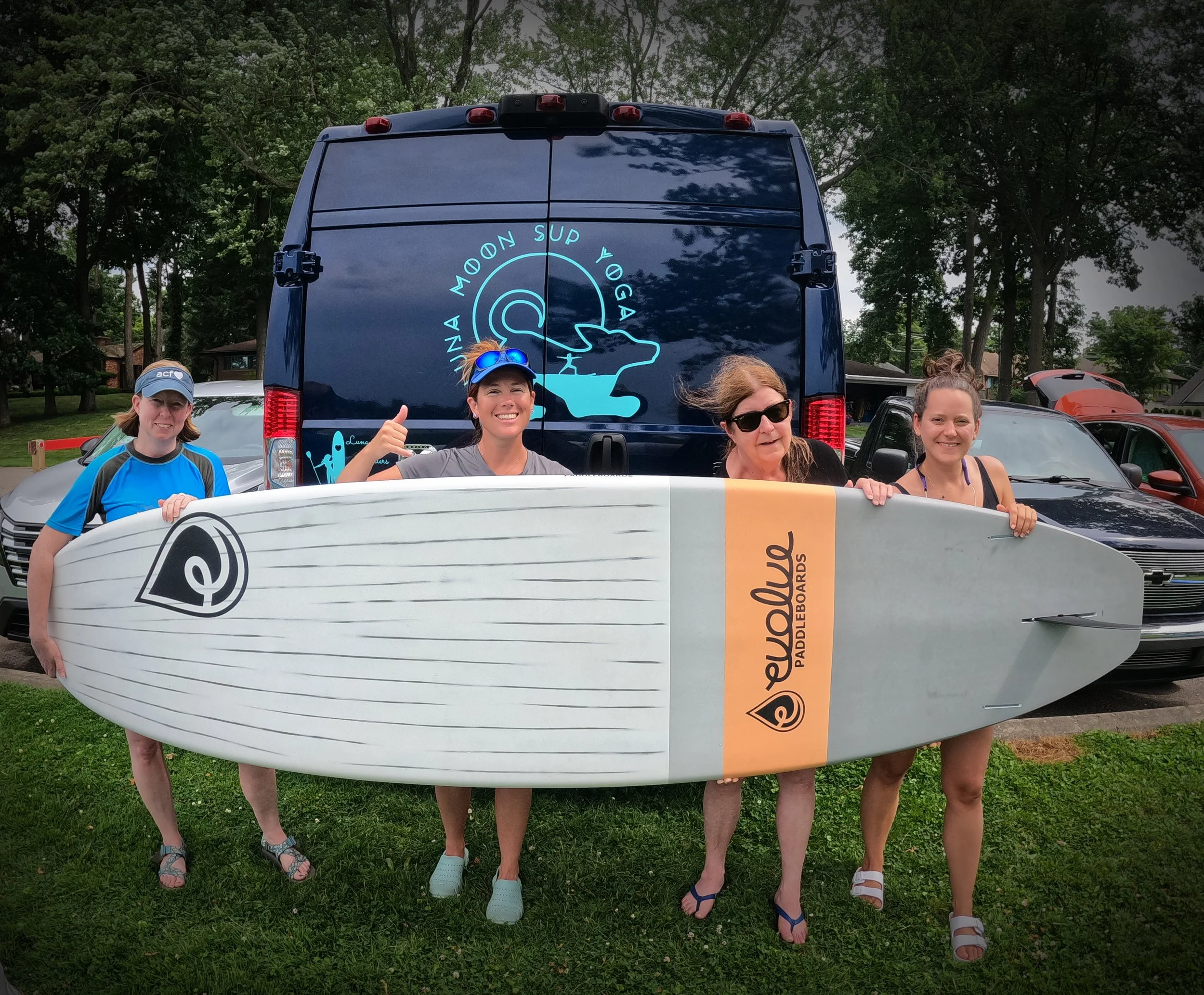 Four women standing outdoors on grass holding a large paddleboard in front of a black van with a logo for Luna Moon SUP Yoga on it. They are smiling, and the woman second from left is giving a thumbs up.