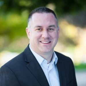 A man in a suit and tie standing outdoors with greenery in the background.