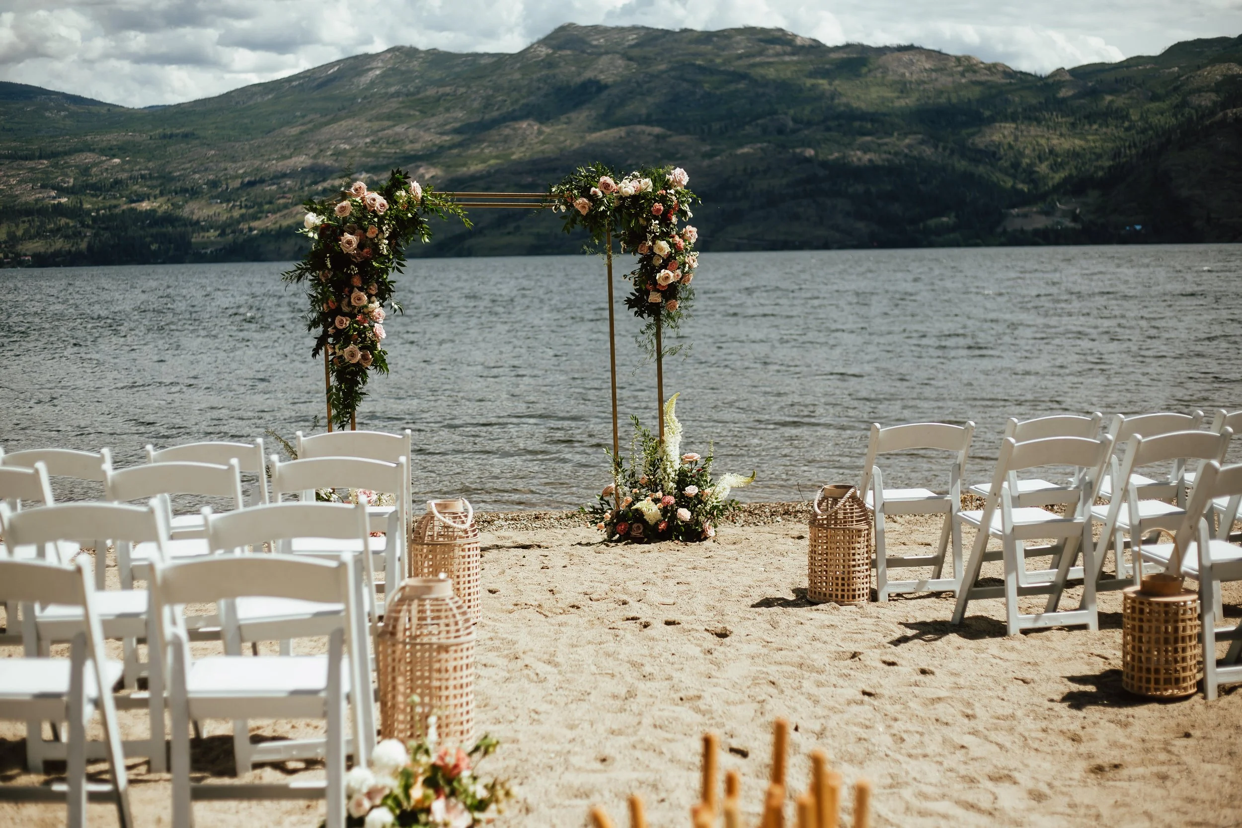 The Cove Lakeside Resort Beach wedding setup with white chairs facing a floral arch by a lake, surrounded by mountains. In West Kelowna BC. Vintage Origami design/decor for the wedding. Day by Knight Events Wedding Planner