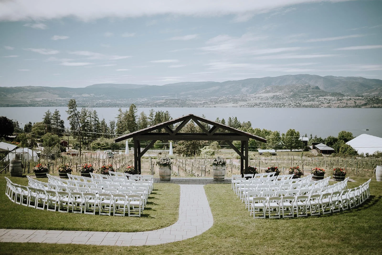 Outdoor wedding ceremony setup with white chairs in rows on a grassy area, facing a wooden pergola , overlooking a lake, vineyards and mountains. Summerhill Pyramid Winery in Kelowna BC. Winery Wedding. Day by Knight Events Wedding Planner.
