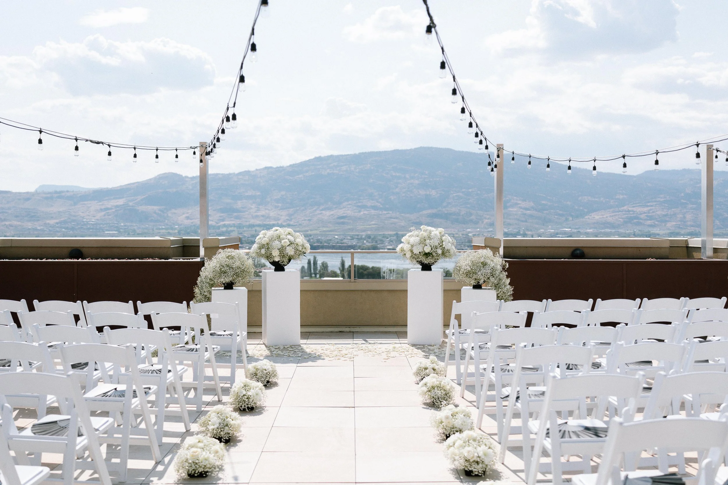 Rooftop Outdoor wedding ceremony setup with white chairs, white floral arrangements, string lights, and a mountain view in the background. Spirit Ridge Resort in Osoyoos BC. Wedding Planner: Day by Knight Events.