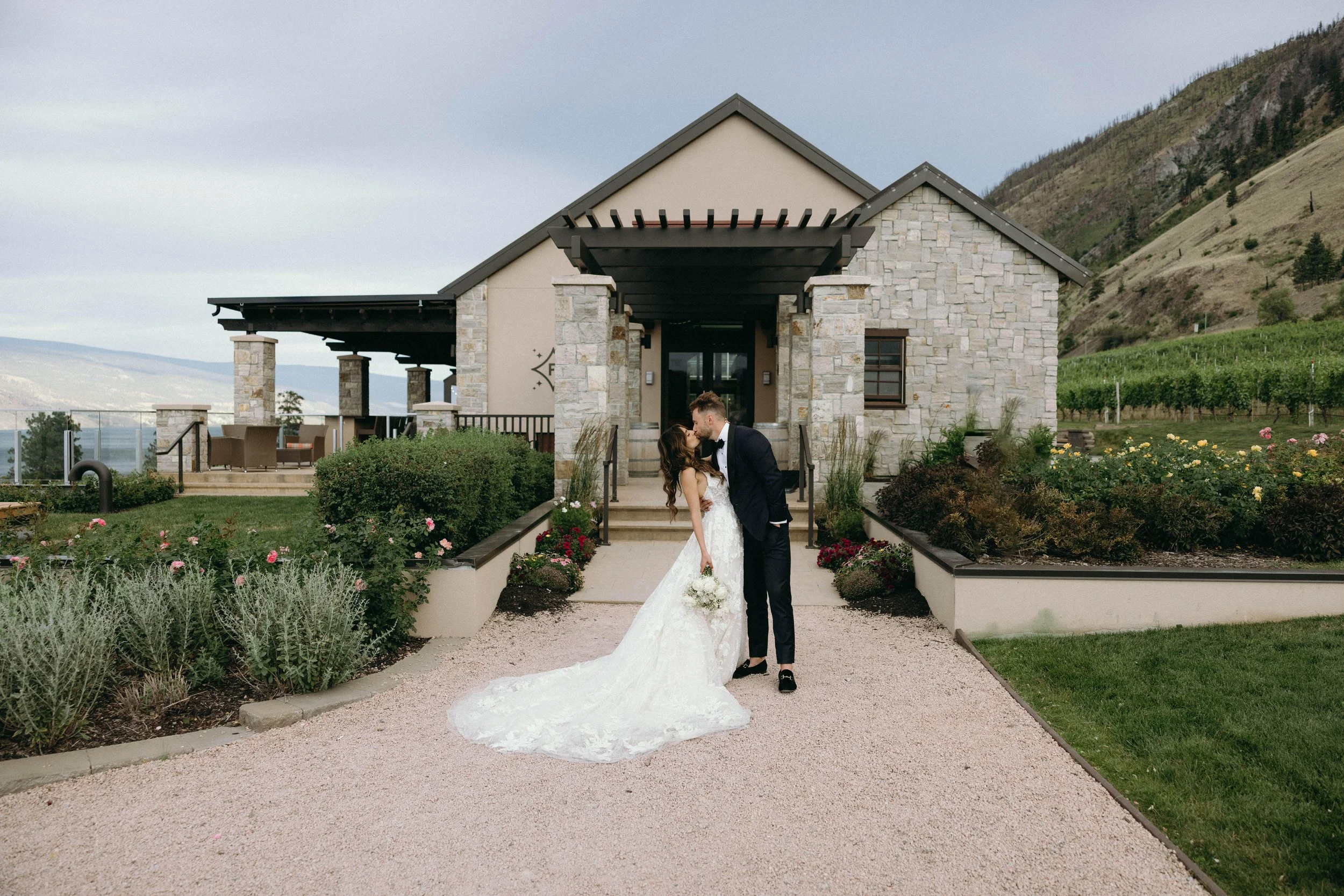 A bride and groom share a kiss in front of a stone and stucco house with a porch, surrounded by a garden and hills in the background.