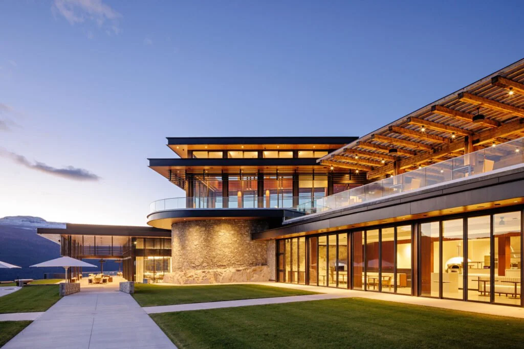 Modern building with large glass windows and wooden accents, located in a scenic area with mountains in the background, during sunset.