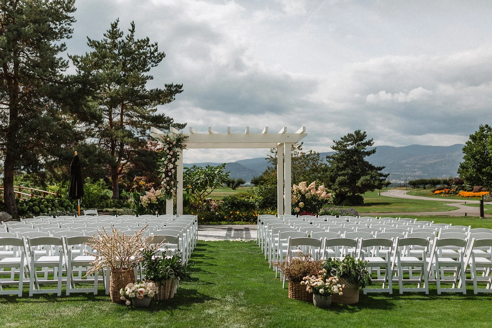Outdoor wedding ceremony setup with white chairs arranged on a grass lawn, decorated with flower arrangements, a white arbor, and scenic mountain view in the background.