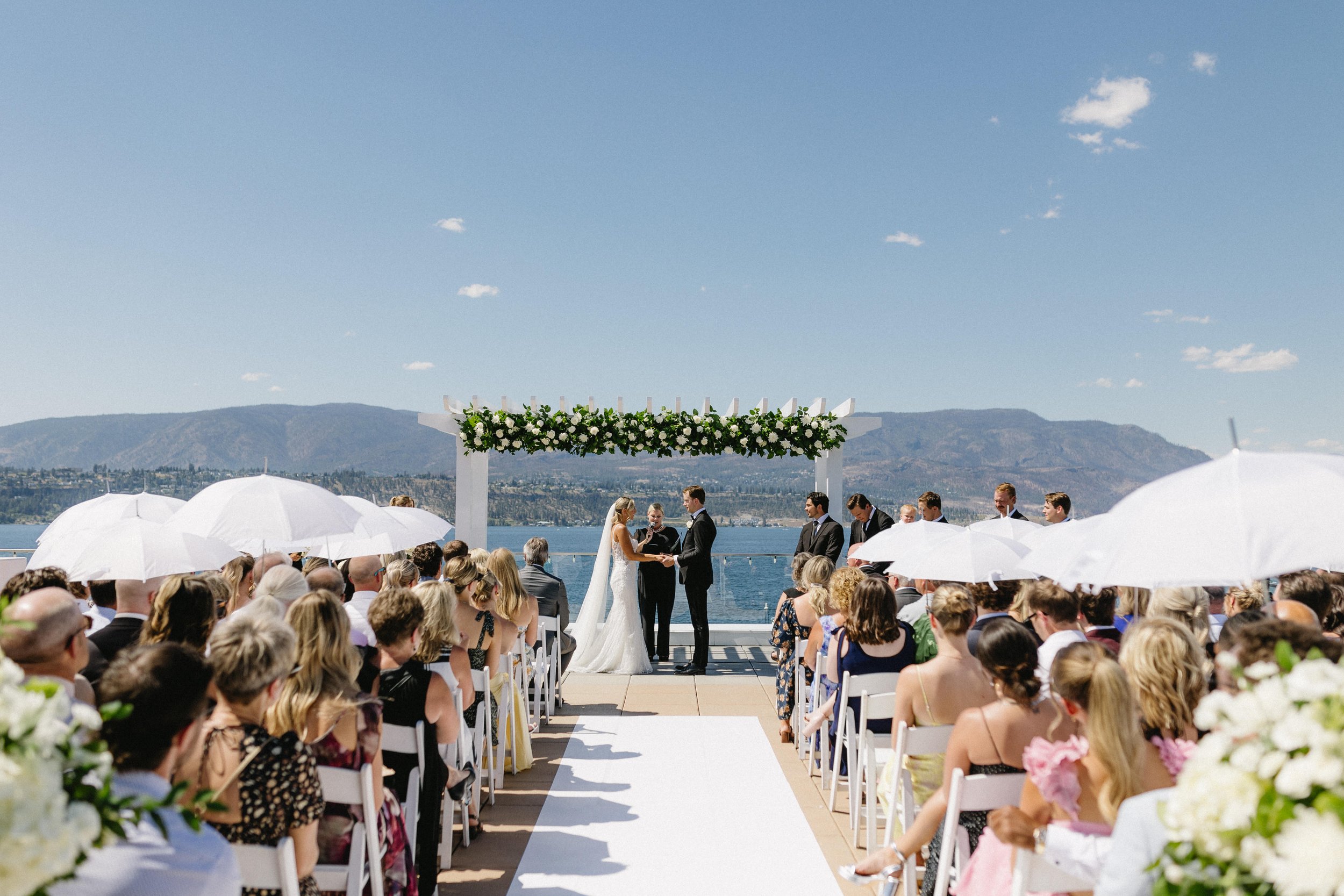 Hotel Eldorado, A wedding ceremony outdoors on a rooftop pato by a lake with a mountain backdrop in Kelowna BC. The bride and groom stand under a decorated arch. Boketto Photography. Day by Knight Events Wedding Planner. NHL Wedding Couple.