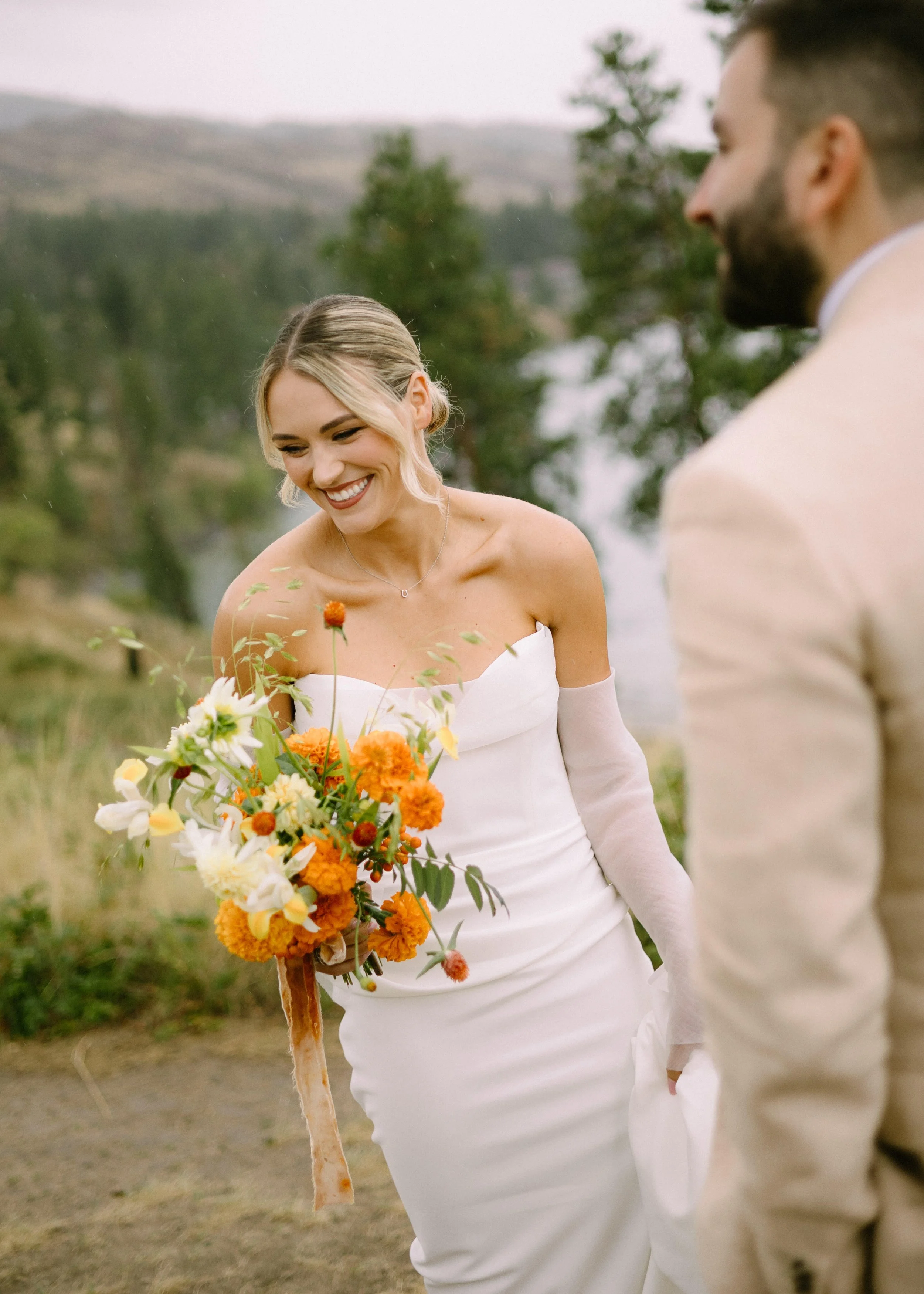A smiling bride in a white strapless wedding dress holding a bouquet of orange, yellow, and white flowers while standing outdoors near a lake and trees.
