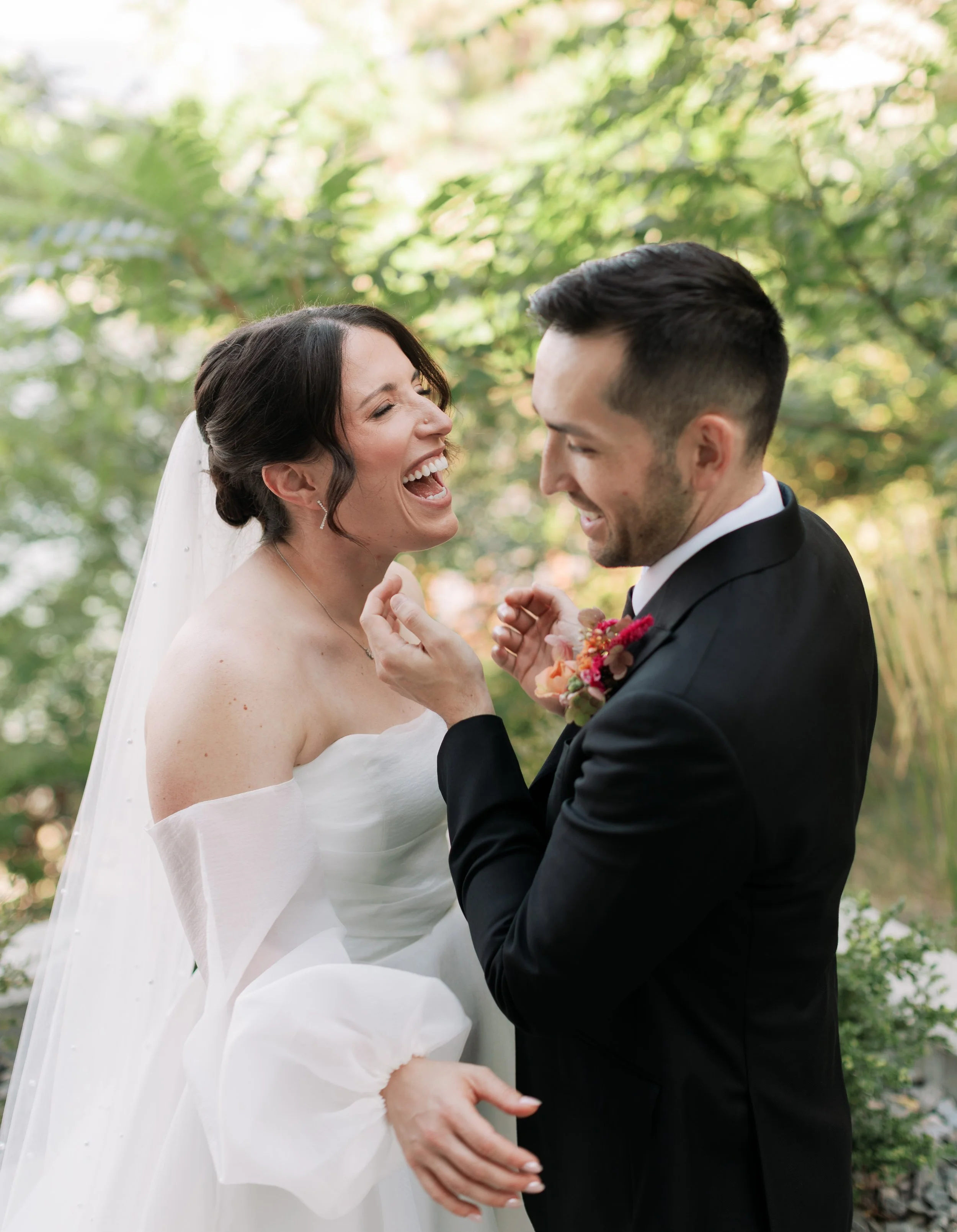 A bride and groom sharing a joyful moment outdoors, with the bride laughing and the groom smiling as he touches her face. The bride wears a white off-the-shoulder wedding dress with sheer sleeves, and the groom wears a black suit with a boutonniere of flowers. Green trees are in the background.