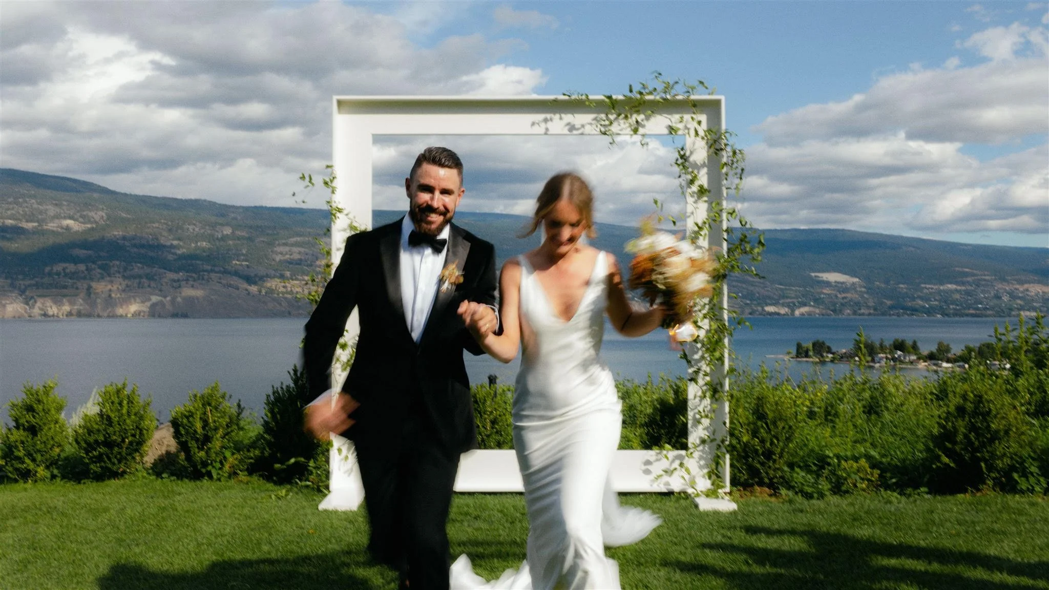 A newlywed couple, a man in a black tuxedo and a woman in a white wedding dress, walking arm-in-arm outdoors on a grassy area near a lake with hills in the background, on a partly cloudy day.