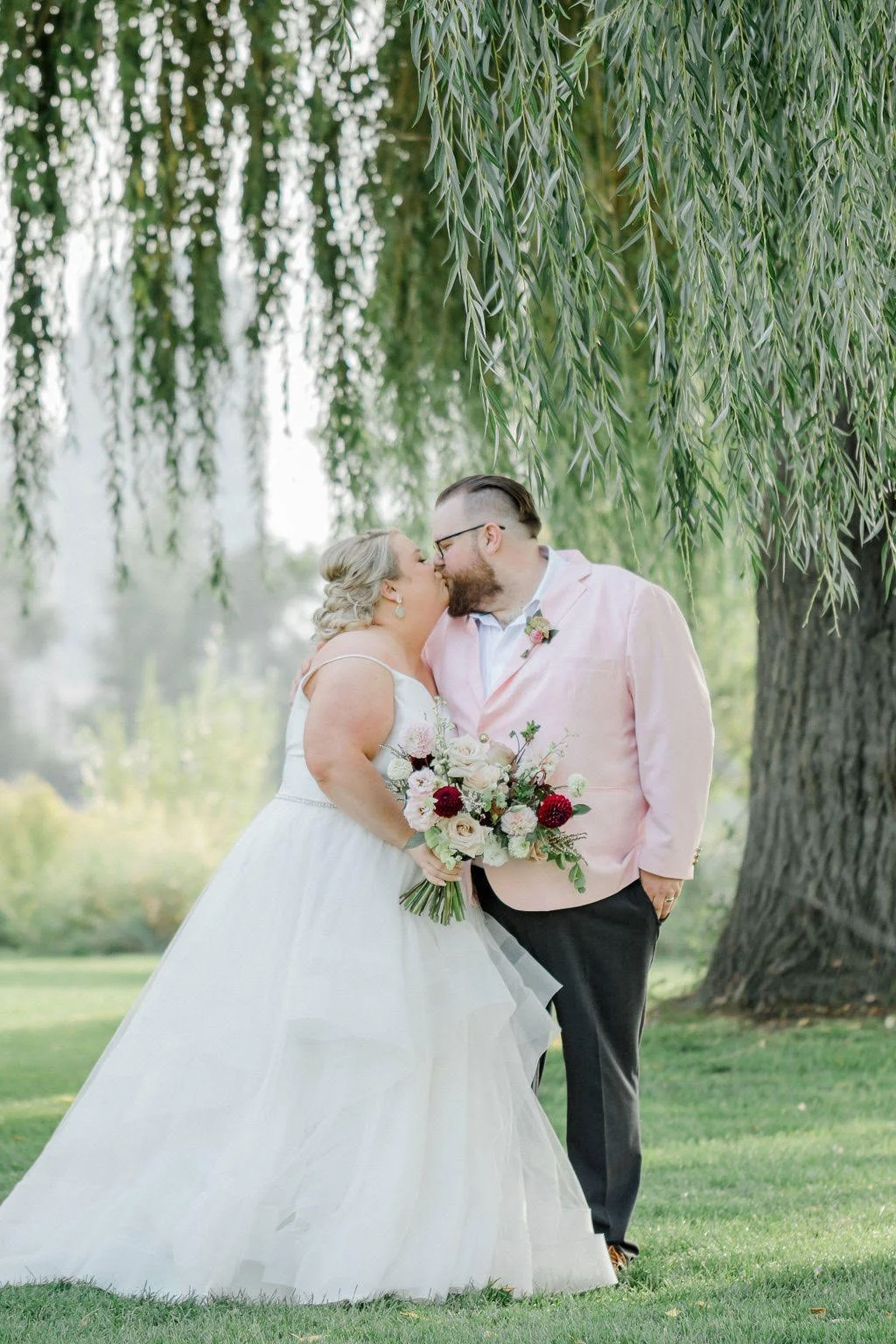 Bride and groom sharing a kiss outdoors beneath a large tree, the bride holding a bouquet of flowers, during their wedding.