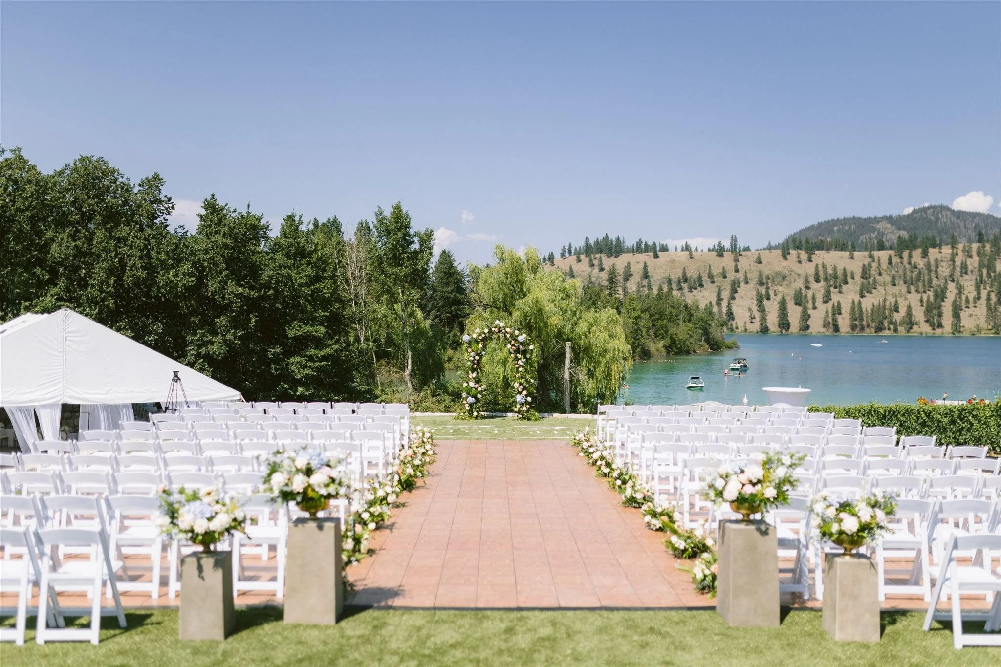 Kelowna BC, Outdoor wedding setup by a lake with white chairs, floral arrangements, and an arch for the ceremony. Day by Knight Events Wedding Planner. Boketto Photography. Private Residence Wedding. Fleurich Creations florals. Avalon Event Rentals.