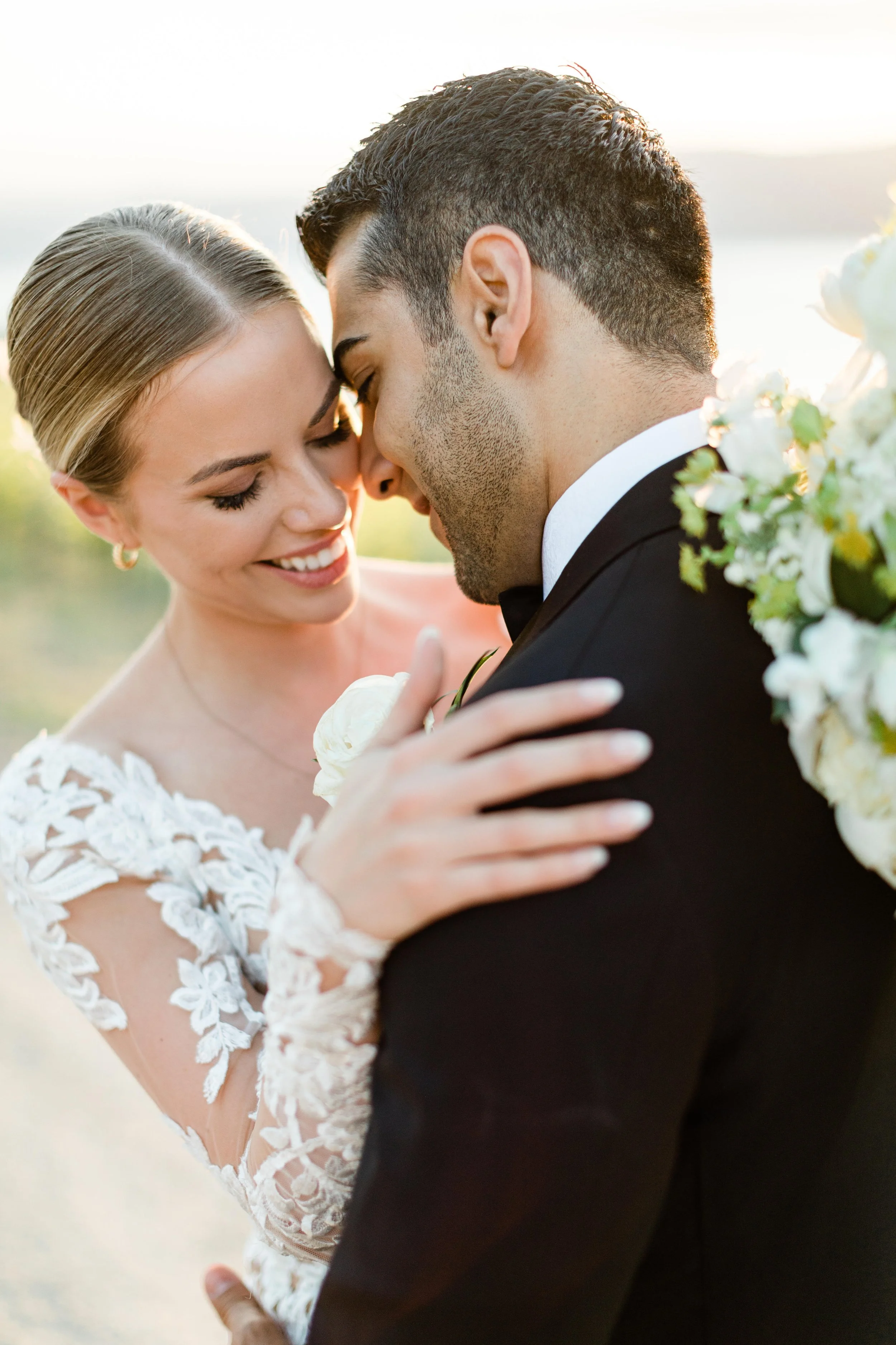 A bride and groom are close together, touching foreheads and smiling, during their wedding outdoors, with a bouquet of white flowers visible.
