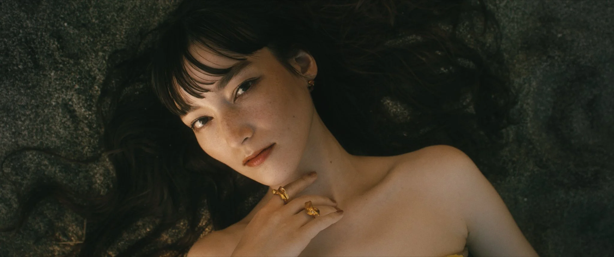 A woman with long dark curly hair lying on a sandy surface, wearing gold jewelry and a yellow top, gazing at the camera.
