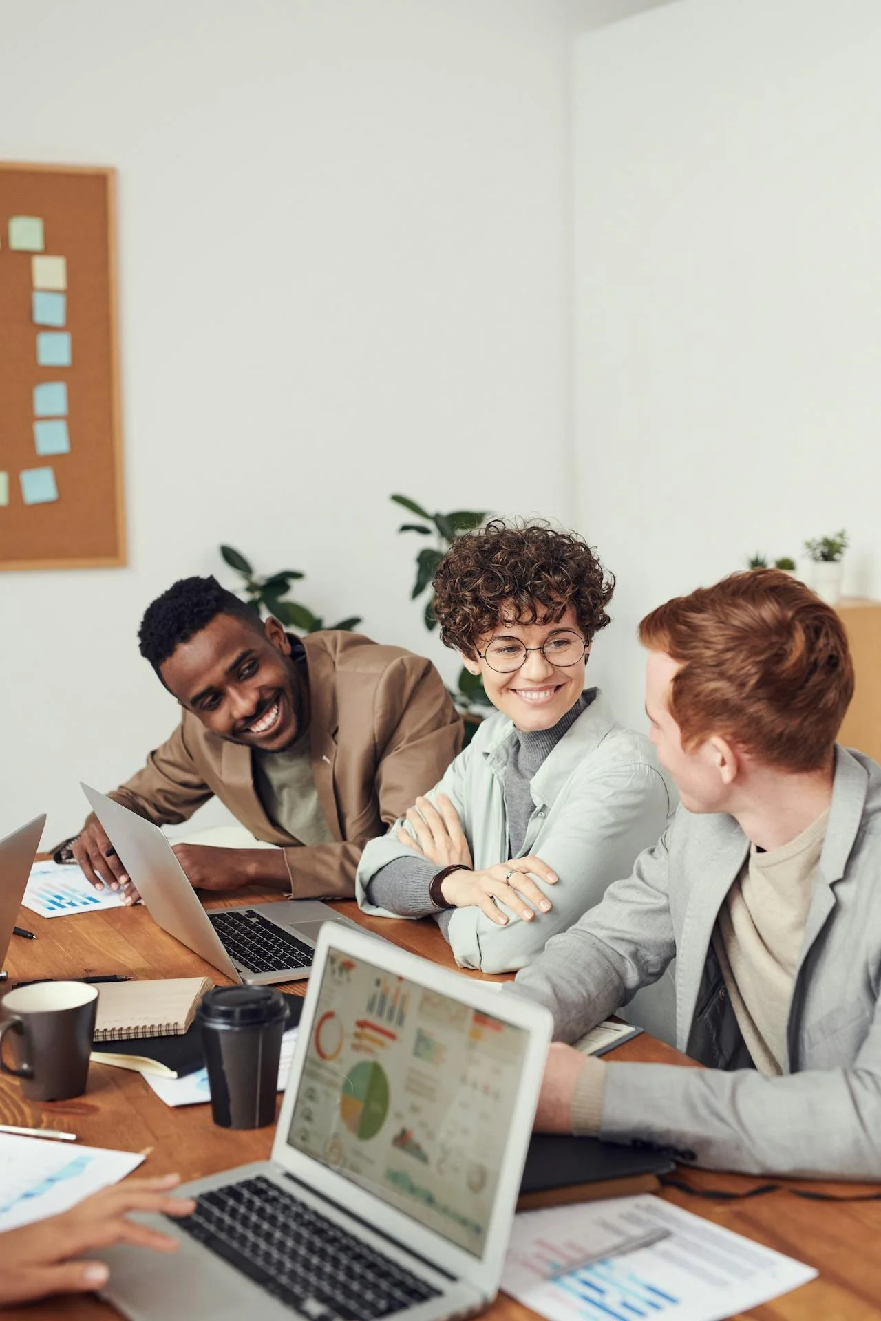 A diverse group of three people sitting at a conference table, smiling and interacting during a meeting with laptops and documents.