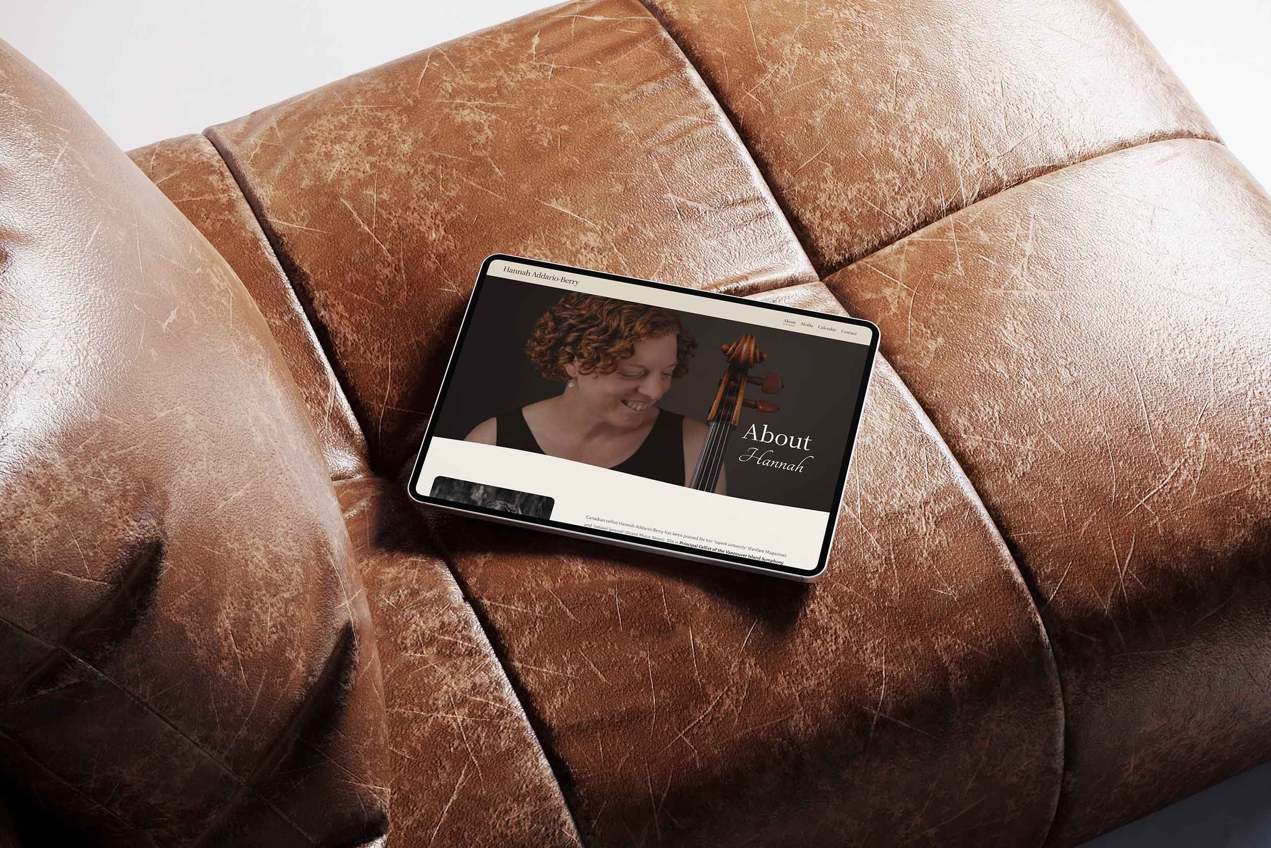 Tablet on a brown leather couch displaying a webpage titled "About Hannah" with a picture of a woman with curly hair and a violin.