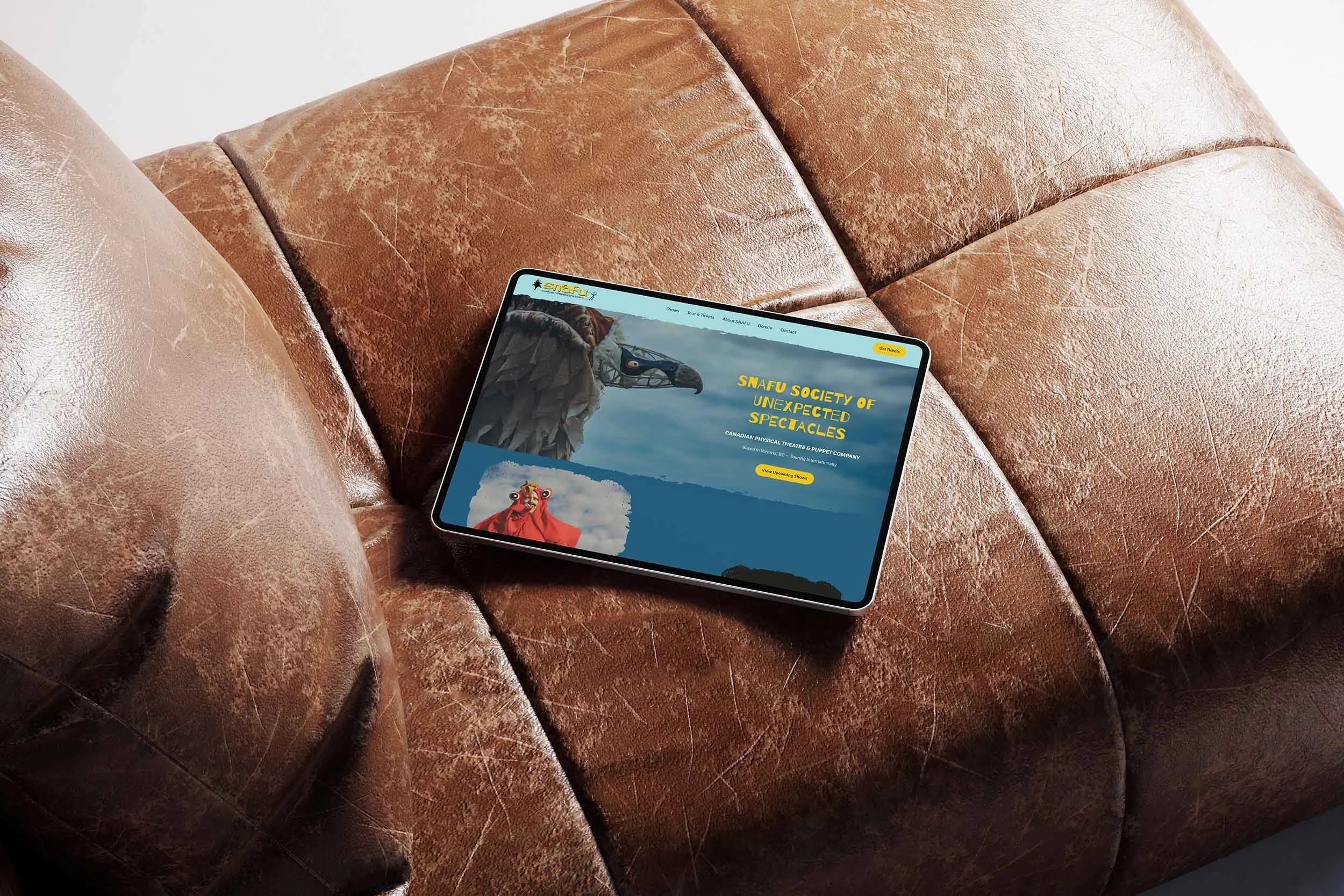 A tablet displaying the SNAFU Society of Unexpected Spectacles website resting on a worn brown leather couch.