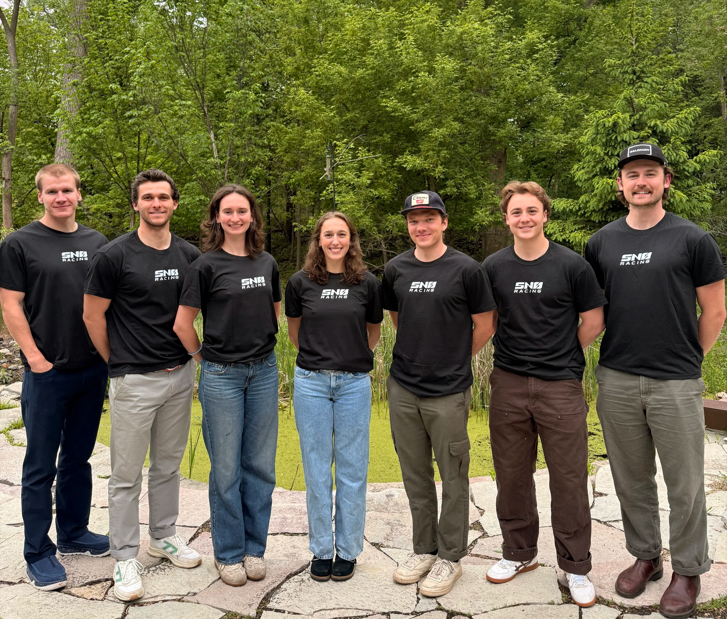 Group of seven young adults standing outdoors in a line against a backdrop of green trees, all wearing matching black T-shirts with white text that says "Snø Racing".