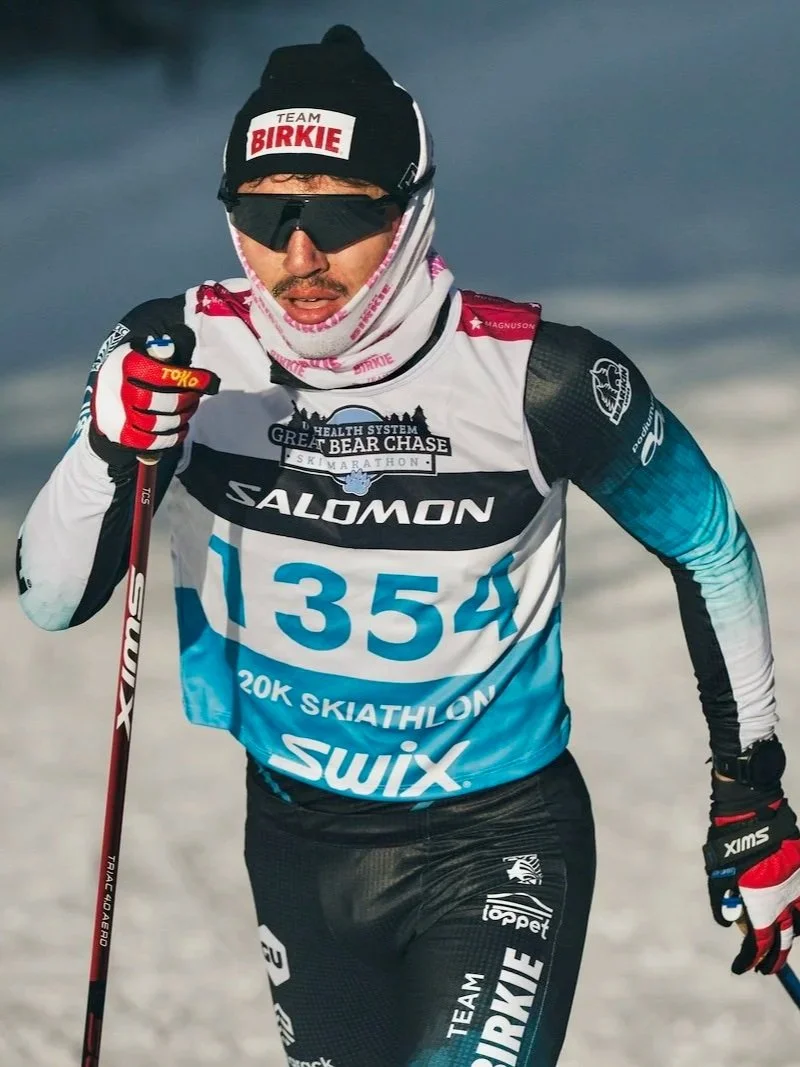 An athlete participating in a winter sports event, dressed in a black, white, and blue outfit with race bib number 1354, wearing sunglasses, a black beanie with a Team Birkie logo, and holding ski poles, skiing on snow.