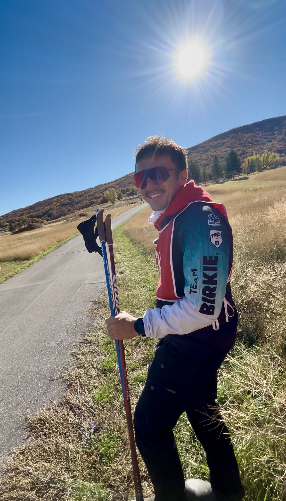 A man dressed in a red, white, and blue sports jacket with sunglasses and a watch standing outdoors on a paved path. He is holding ski poles and smiling at the camera. The background features grassy hills, trees, and a bright sun in a clear blue sky.