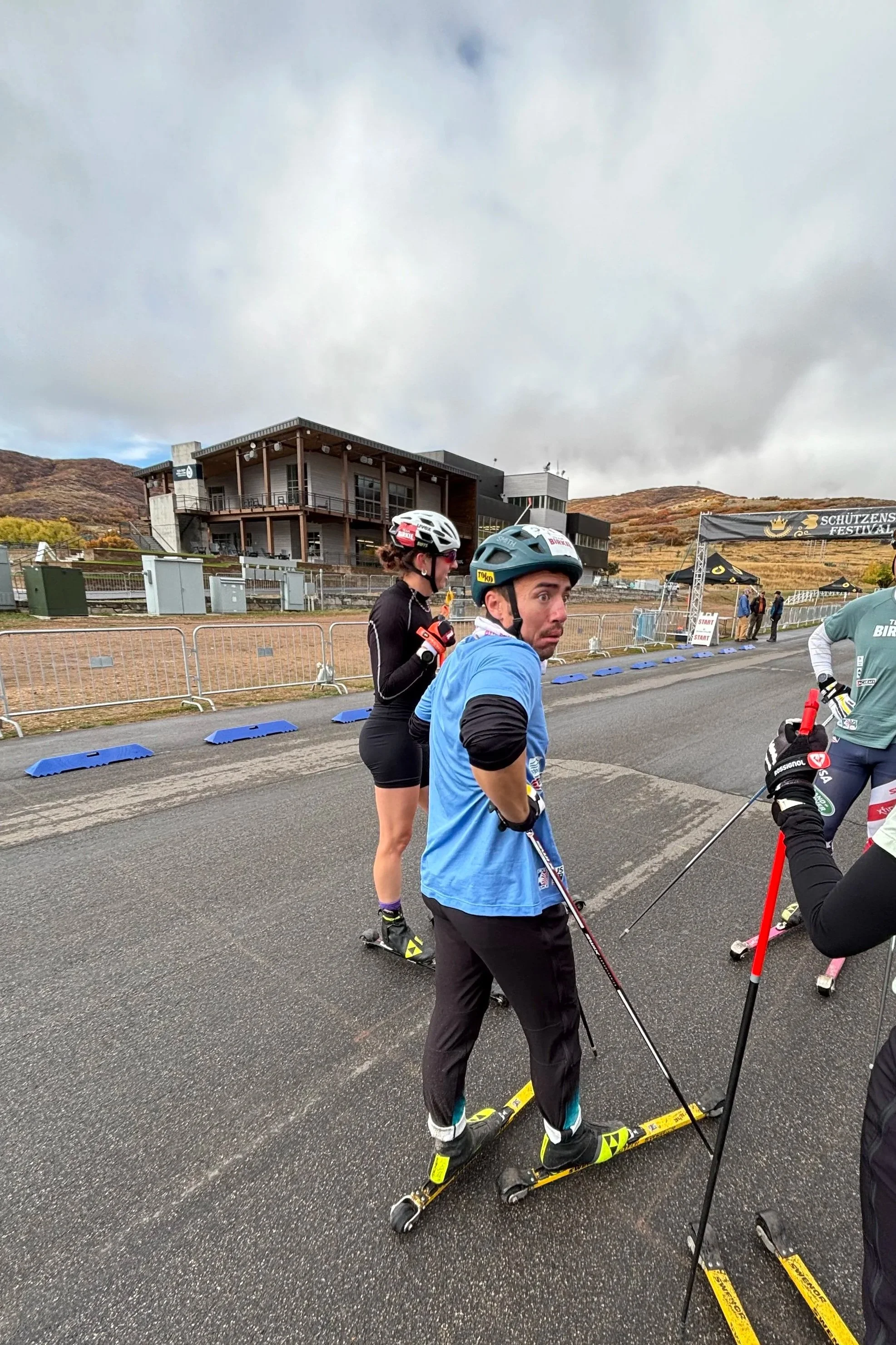 People dressed in athletic gear and helmets preparing for roller skiing at an outdoor event with a banner, fencing, and buildings in the background.