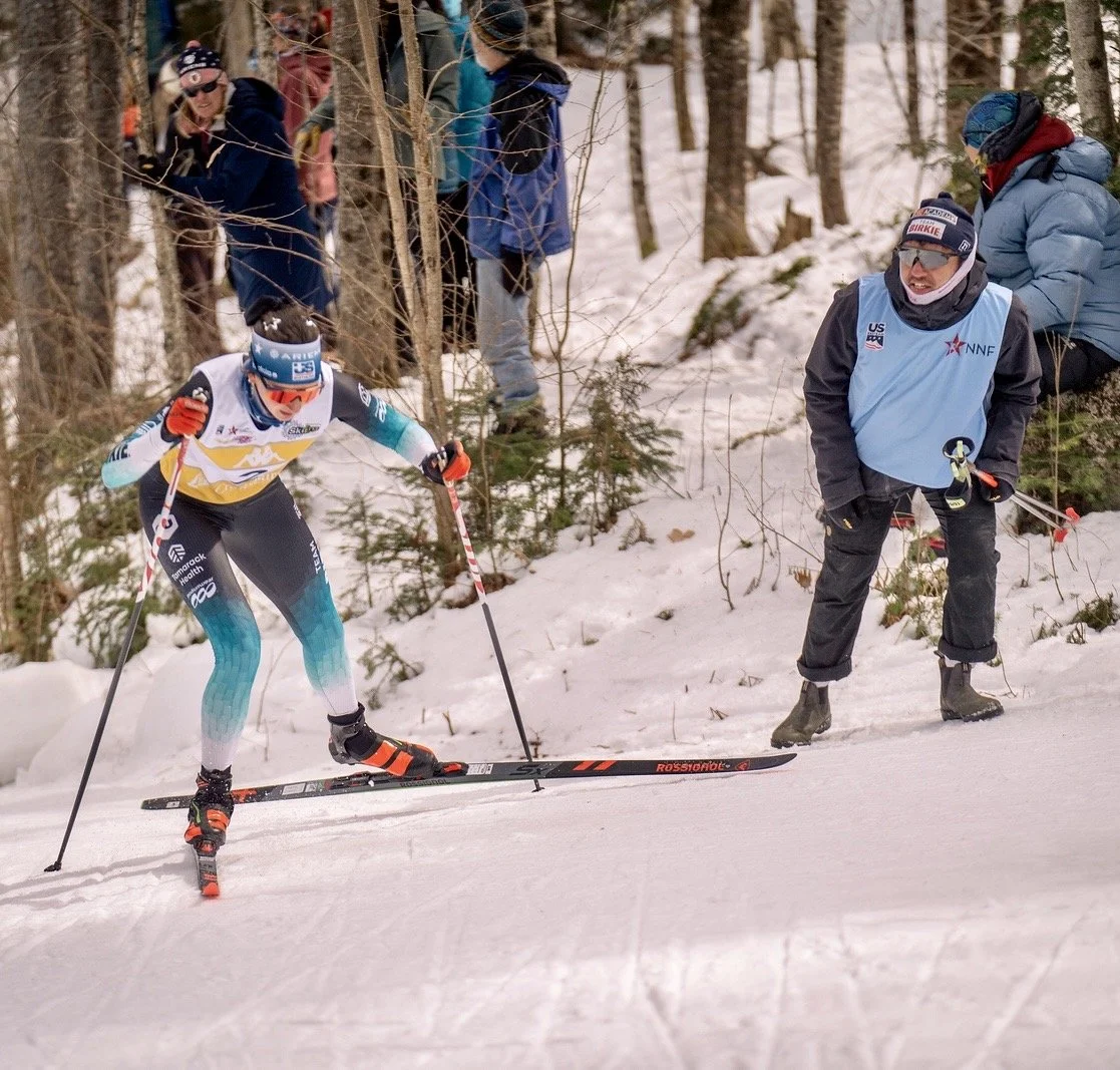 A female cross-country skier in a colorful uniform racing on snow, with spectators and a coach watching in the wooded background.
