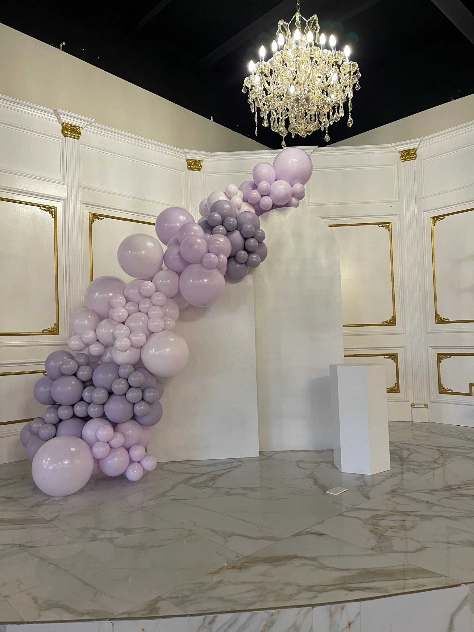 Decorative display with a cascade of purple and lavender balloons on a white backdrop in a luxurious room with a chandelier.