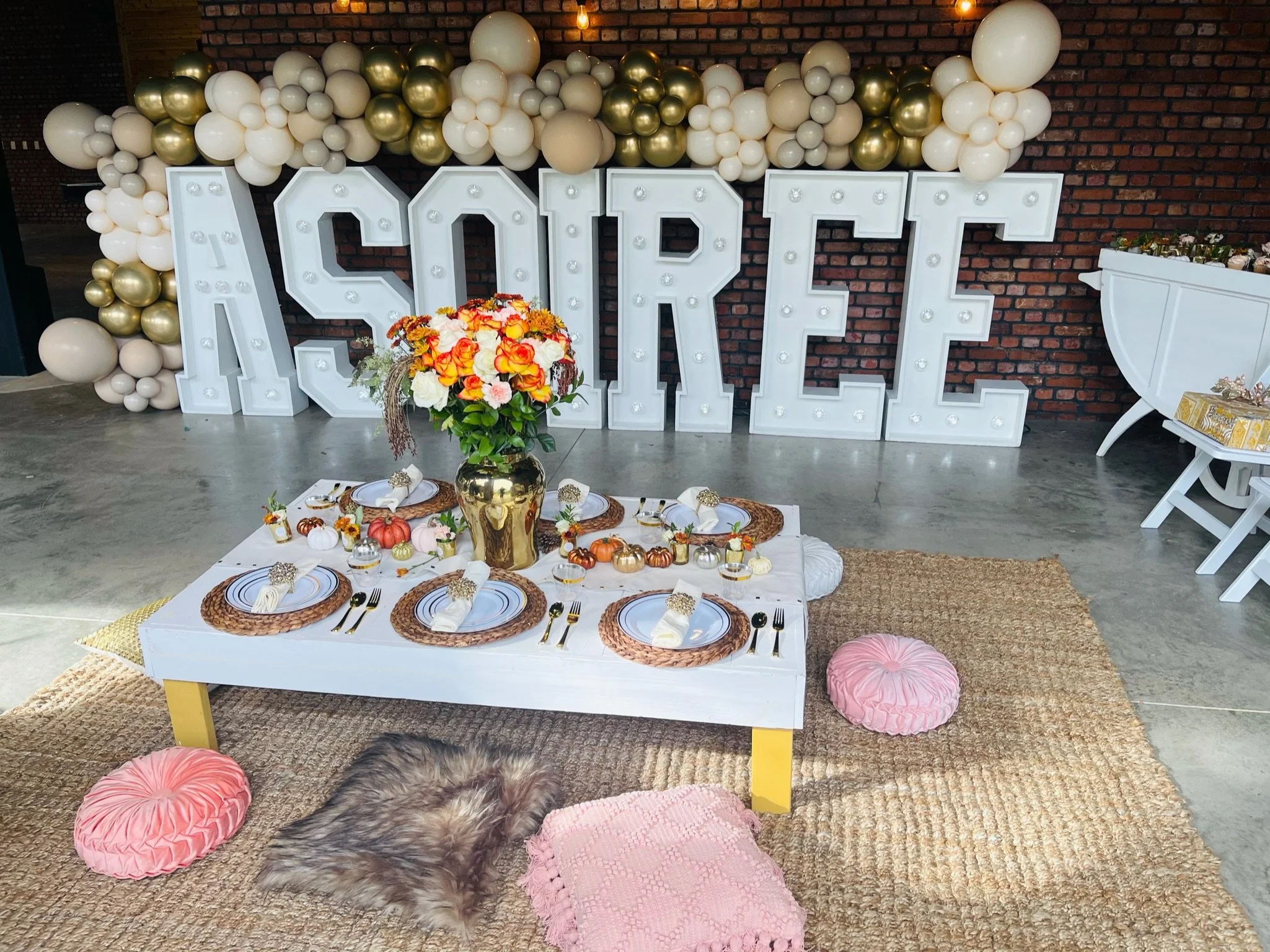 Decorative indoor setup for a celebration featuring a large white sign spelling 'AOREE' adorned with bulbs and balloons in gold, white, and cream colors. A low table set for four, with gold-rimmed plates, cutlery, and cloth napkins, is decorated with
