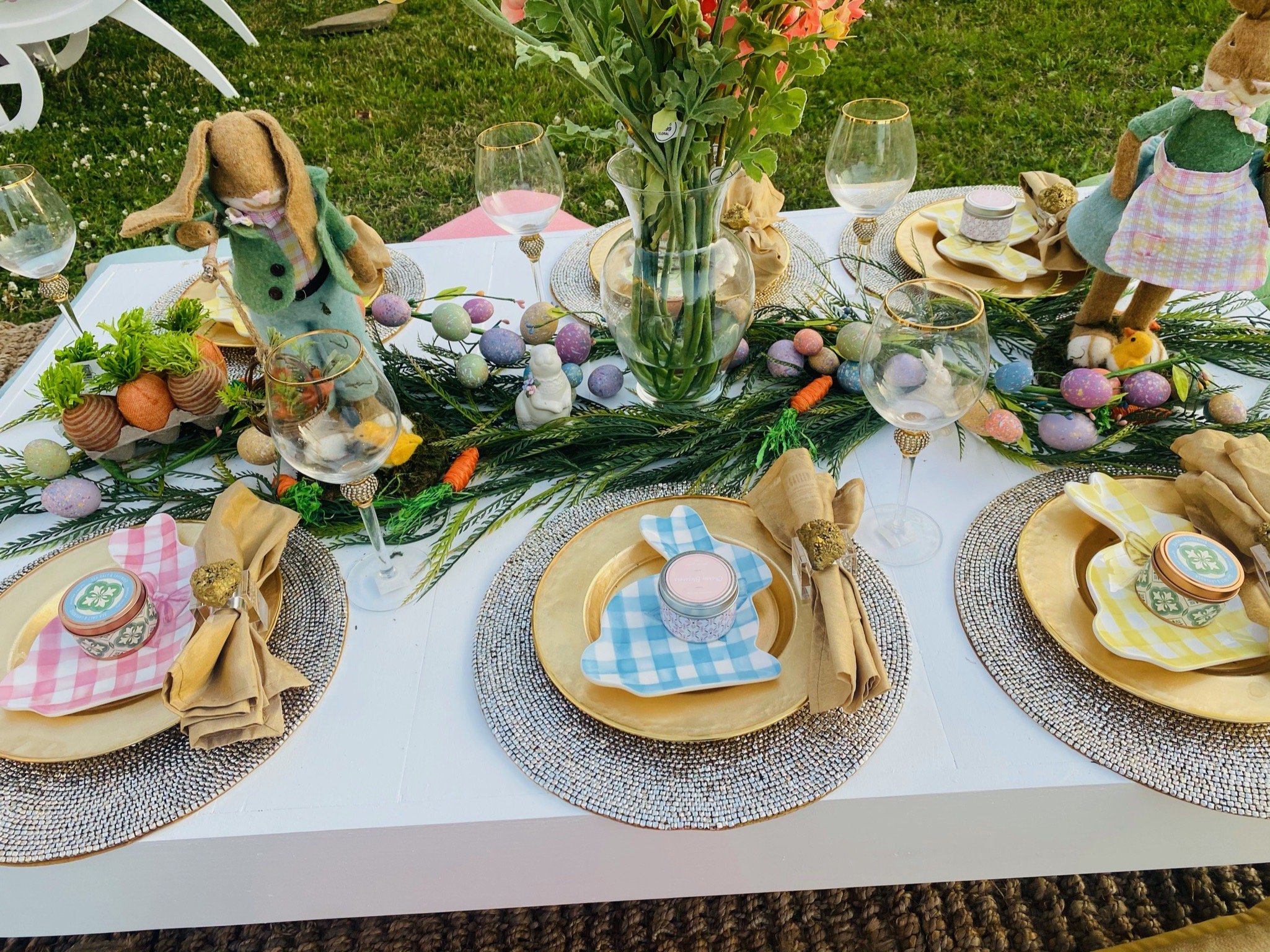 Easter table setting with decorated plates, napkins, glassware, spring-themed decorations, plush bunny and chick figurines, and a centerpiece of flowers and colorful eggs.