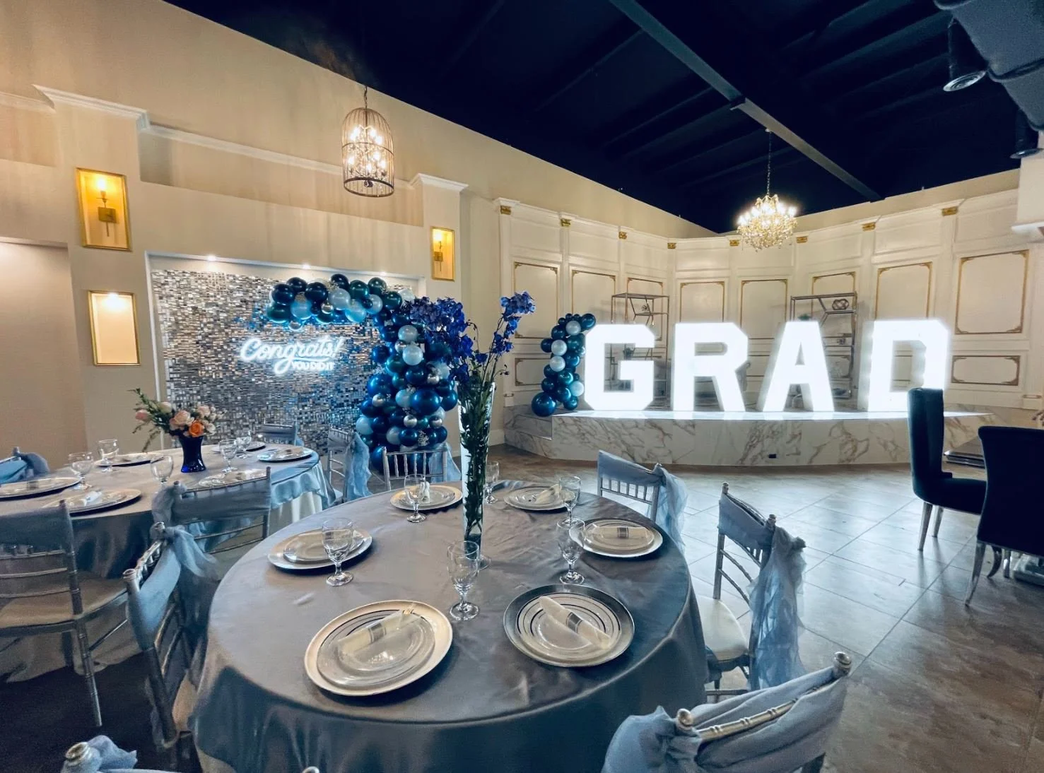 Decorated reception area for a graduation celebration with a large illuminated "GRAD" sign, balloon arrangements in blue, silver, and white, a pastel-colored backdrop with a neon sign reading "congrats graduate," and tables set with white tablecloths