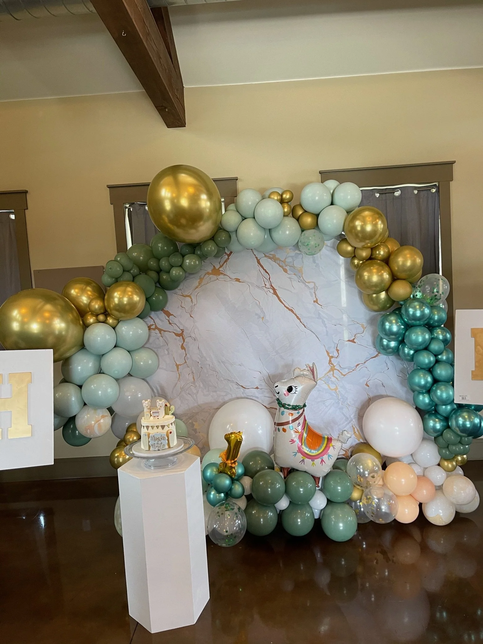 Balloon arch with gold, green, teal, white, and clear balloons, a marble-patterned backdrop, a llama balloon, and a small cake with a gold number one, for a celebration.