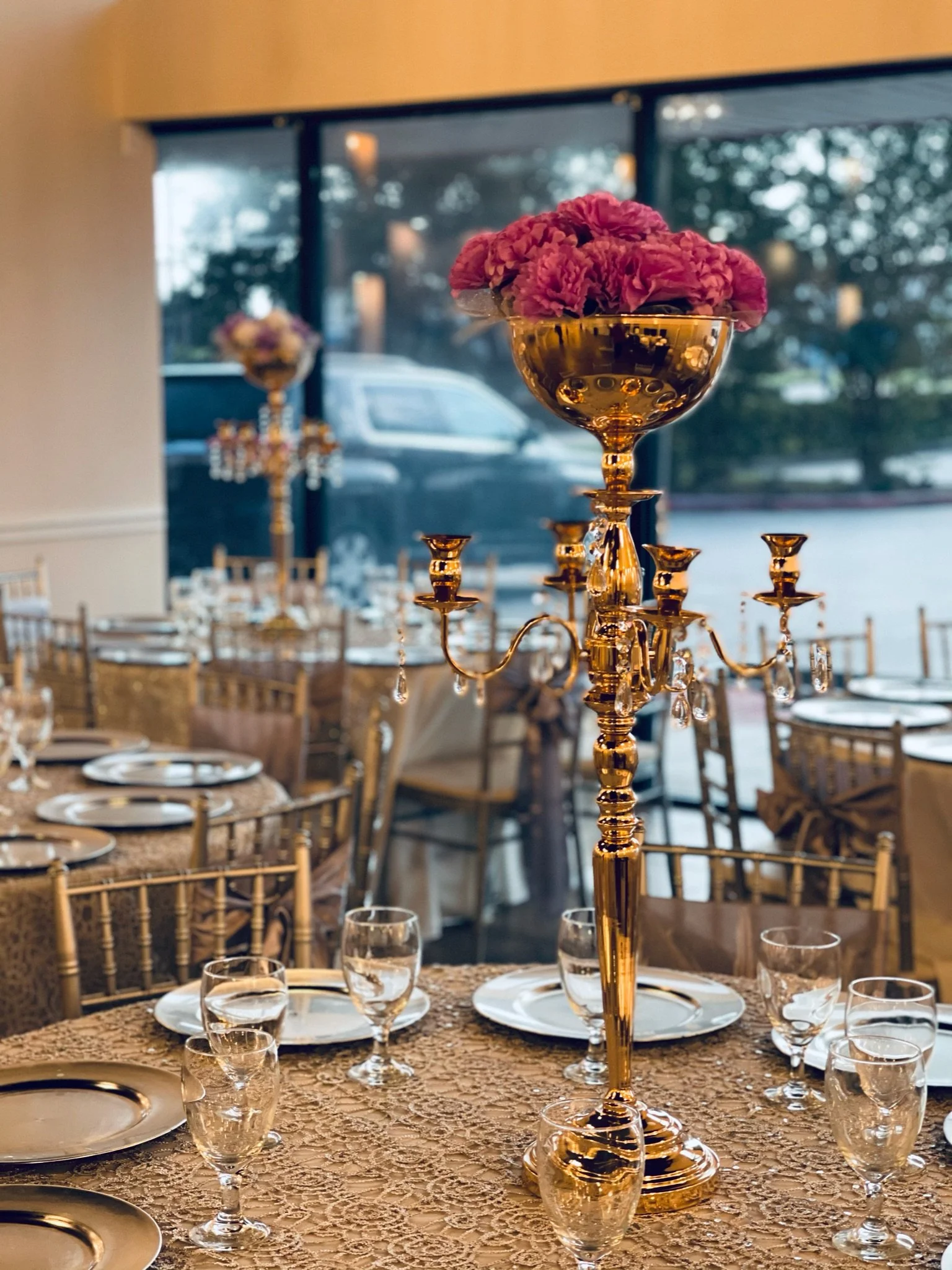 Elegant dining table decorated with a gold candelabrum topped with pink flowers, set with white plates, wine glasses, and cutlery in a well-lit reception hall.