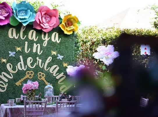 Decorative outdoor scene with a green backdrop with large paper flowers, gold lettering that reads 'aloha in Wonderland,' and small butterfly decorations. There are tables and chairs set up, with floral centerpieces, and a white teddy bear. The setti