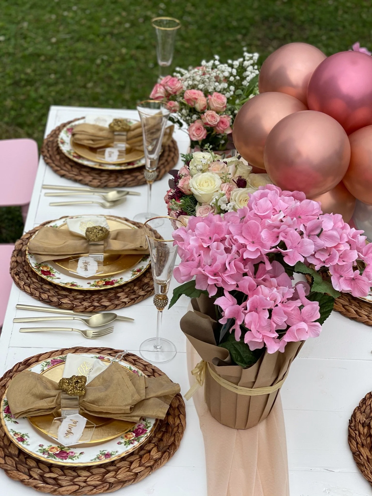 A decorated outdoor dining table with pink and gold balloons, pink and white flowers, gold flatware, floral plates, and gold napkin rings.