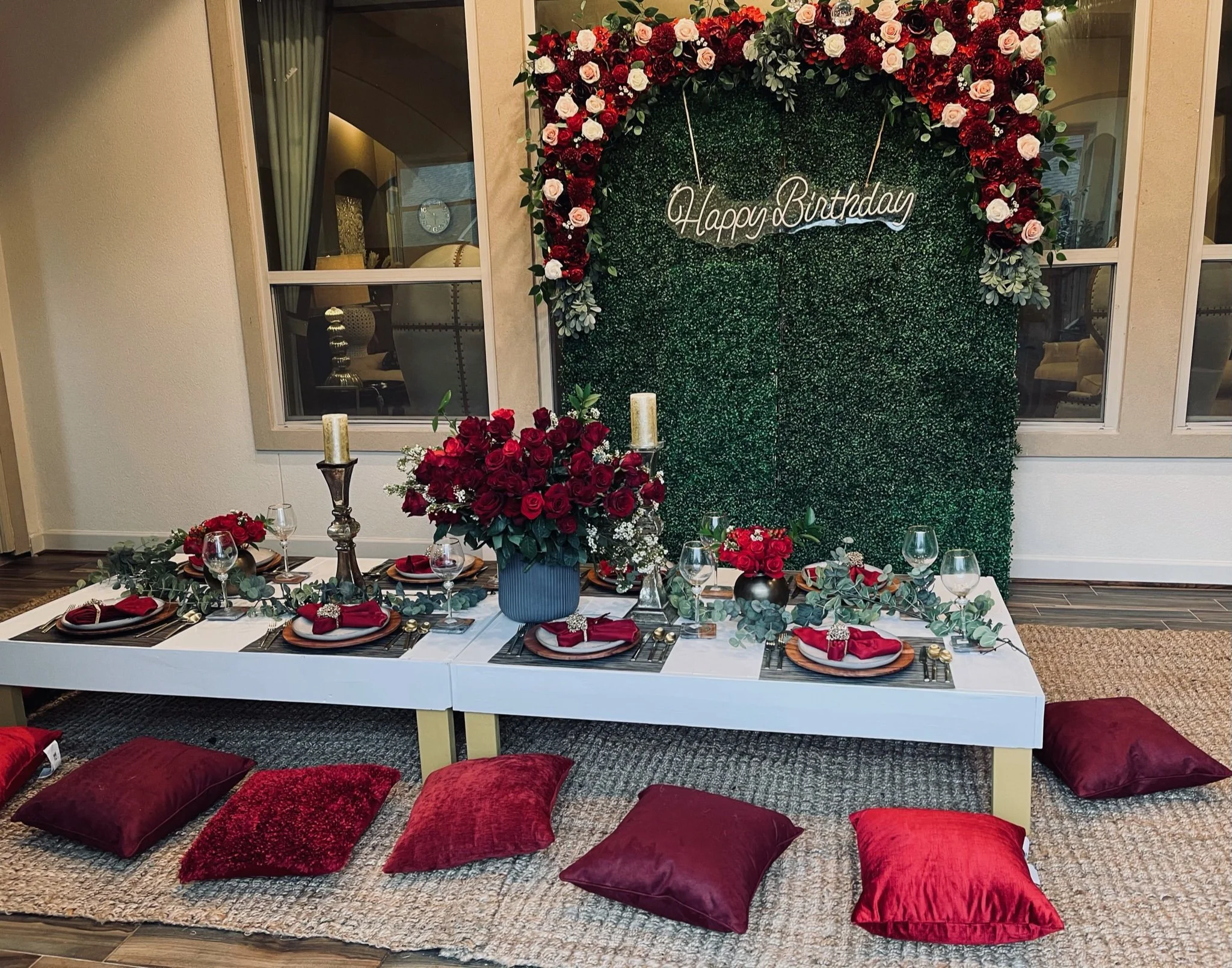 A low table decorated for a birthday celebration with red and white flowers, candles, and wine glasses. The table is surrounded by red cushions on a carpeted floor. Behind the table is a green hedge wall with a pink and white floral arch and a neon s