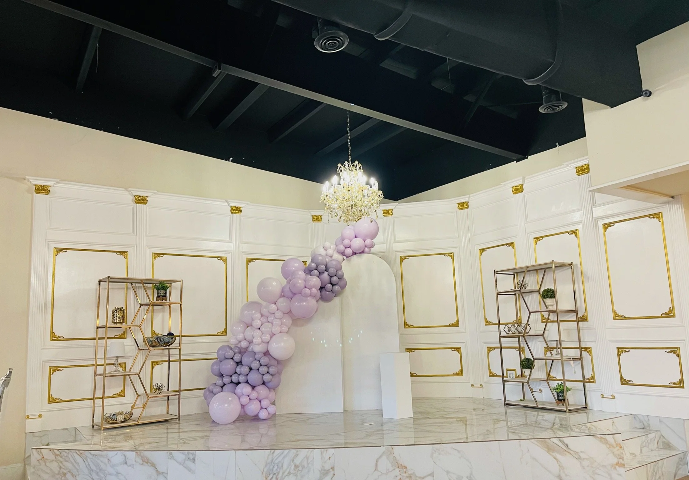 Interior space with white walls decorated with gold trim, featuring a floating balloon arrangement in shades of pink and purple, a chandelier hanging from a black ceiling, and two gold shelving units with decorative items and potted plants on a marbl