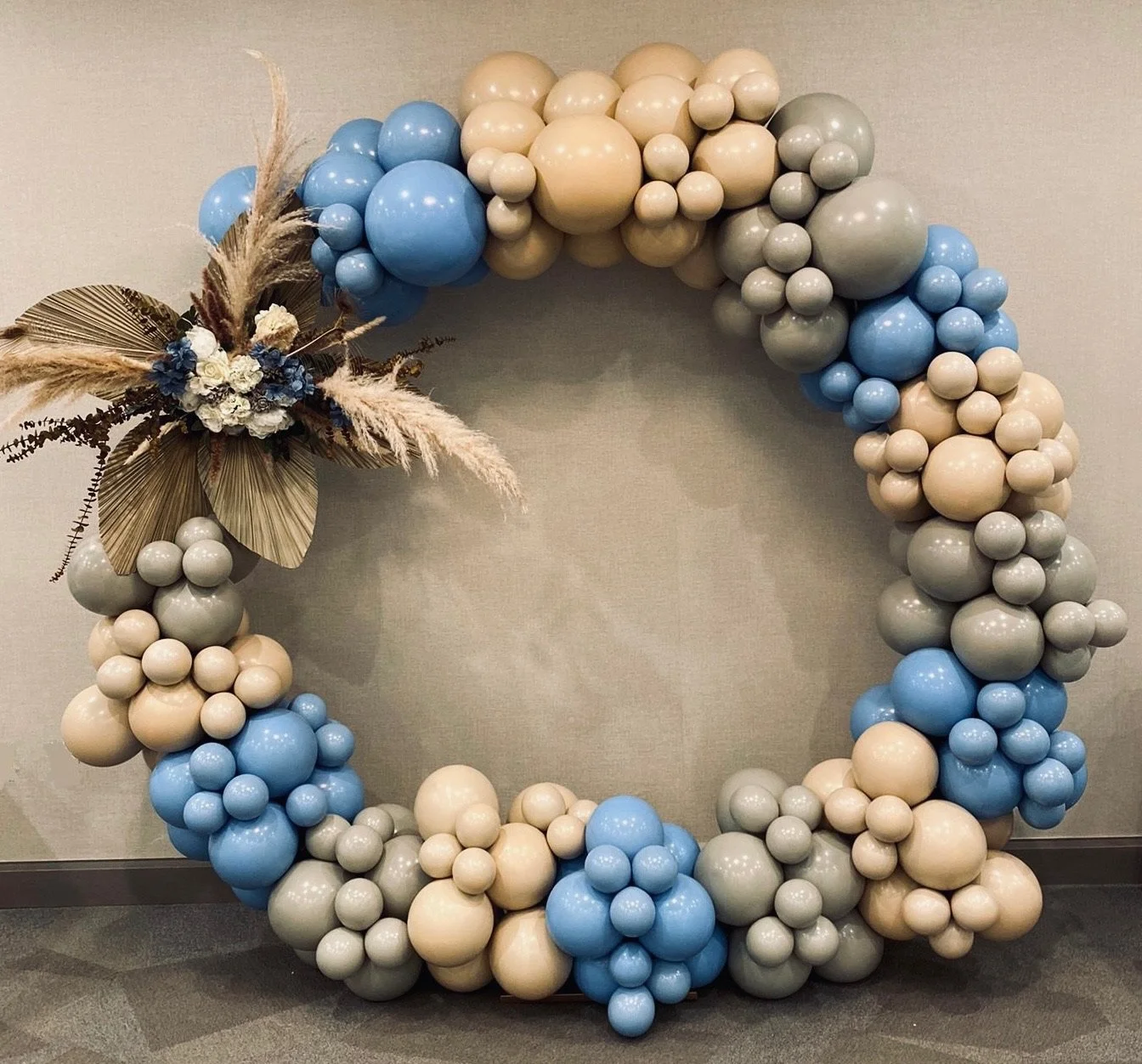Decorative balloon wreath with beige, gray, blue, and cream-colored balloons, adorned with dried and faux floral accents and pampas grass.