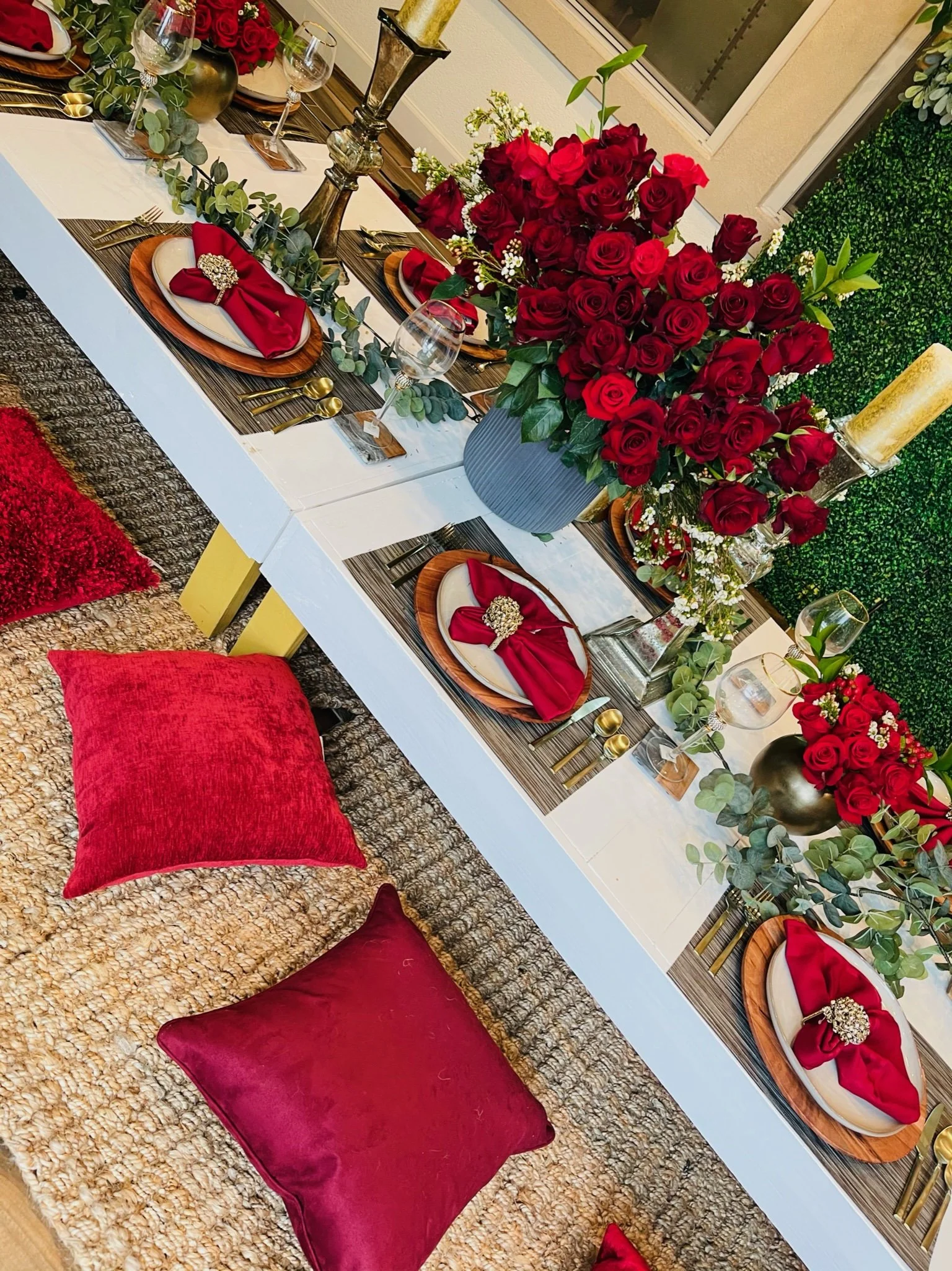 A beautifully decorated dining table with red roses in a large vase as the centerpiece, set with plates, red napkins with brooches, gold utensils, wine glasses, and candles. Cushions are placed on the floor around the table, creating a cozy seating a