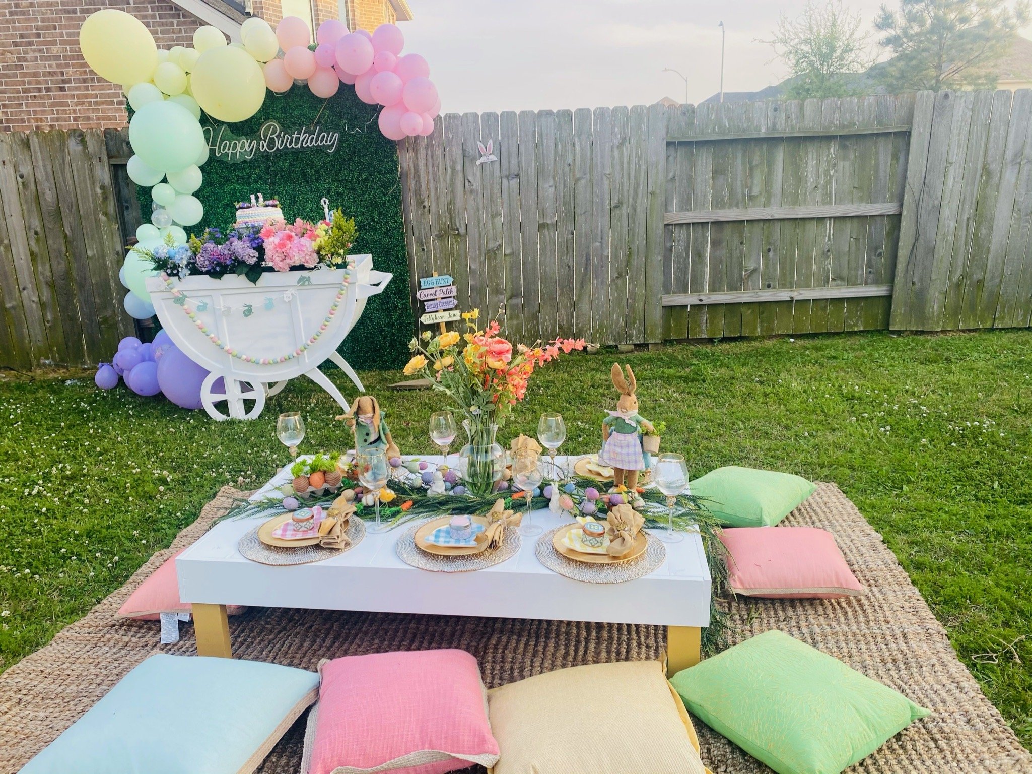 Backyard birthday celebration setup with a low table on a woven rug, surrounded by colorful cushions, decorated with flowers, bunny figurines, and tableware. In the background, a decorative green wall with a 'Happy Birthday' sign, balloons in pastel 