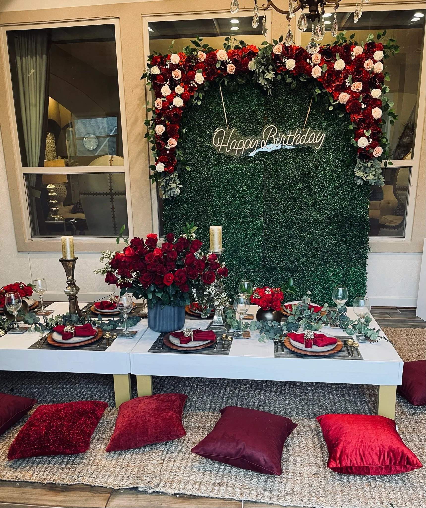 A birthday celebration setup with a green hedge backdrop decorated with a heart-shaped floral arrangement of red, white, and pink roses, and a neon sign that reads 'Happy Birthday.' A white low table in front is set with plates, red napkins, glasses,