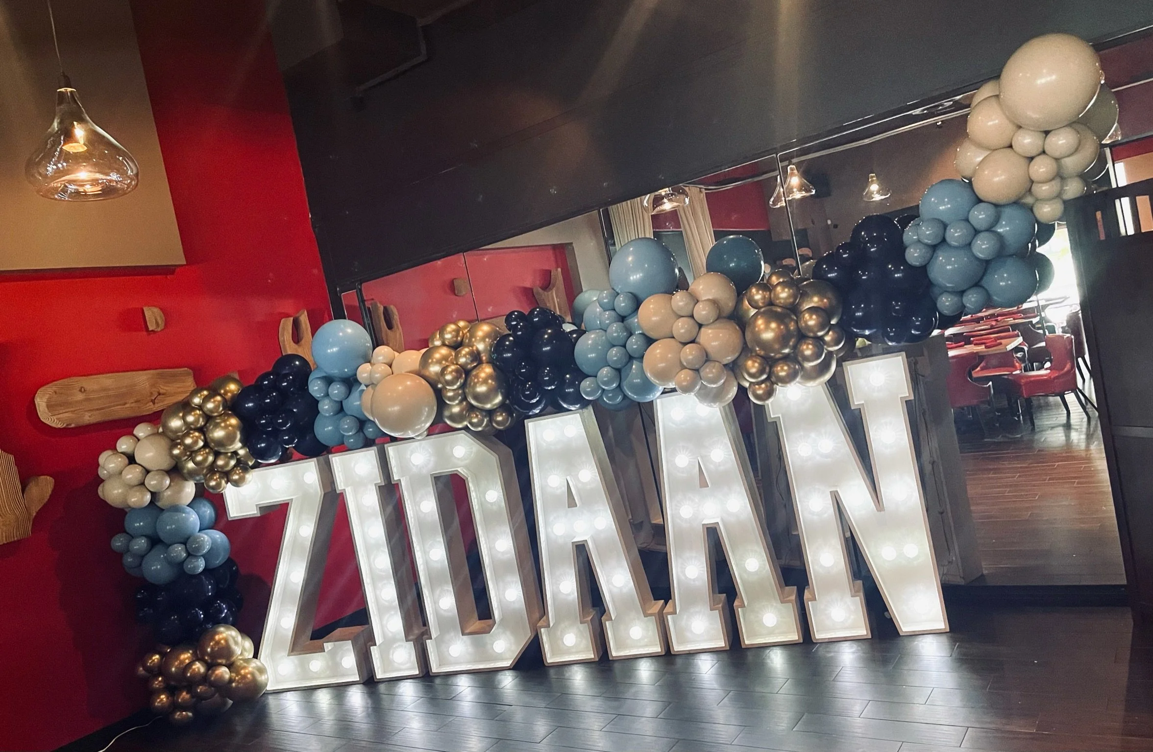 Decorative sign spelling 'ADAM' with large illuminated letters, surrounded by a colorful balloon arch in pastel blue, white, gold, and black, in a decorated indoor event space.