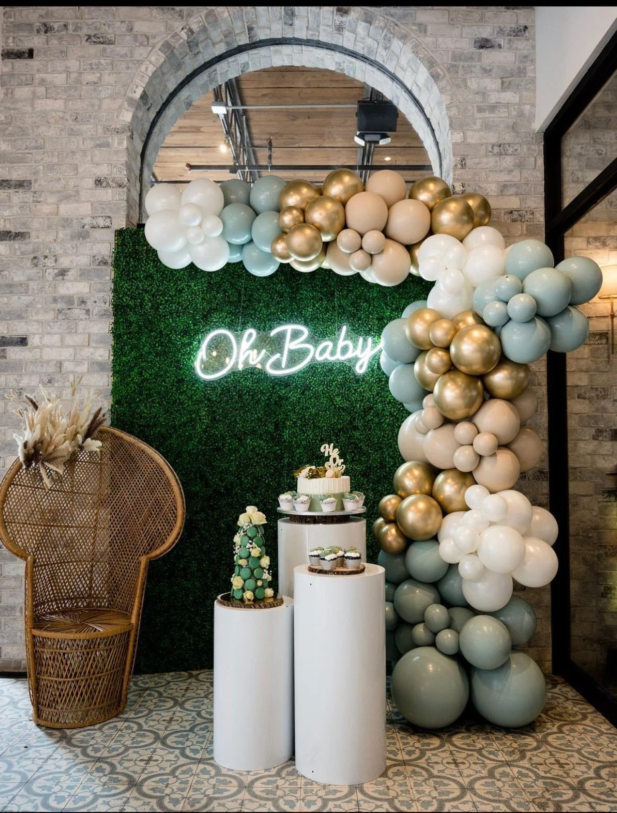 Decorative setup with a green leafy backdrop, a neon sign that reads 'Oh Baby', a balloon arch in white, gold, and light blue, a wicker chair with dried flowers, and three white cylindrical pedestals displaying cupcakes and a small decorated cake for