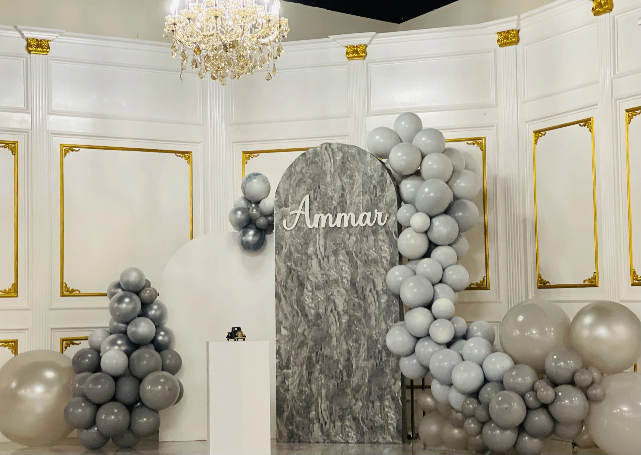 Elegant event backdrop with a gray marble panel, white and gray balloons, golden ornate wall details, and a chandelier, with the name "Ammar" displayed on the marble panel.
