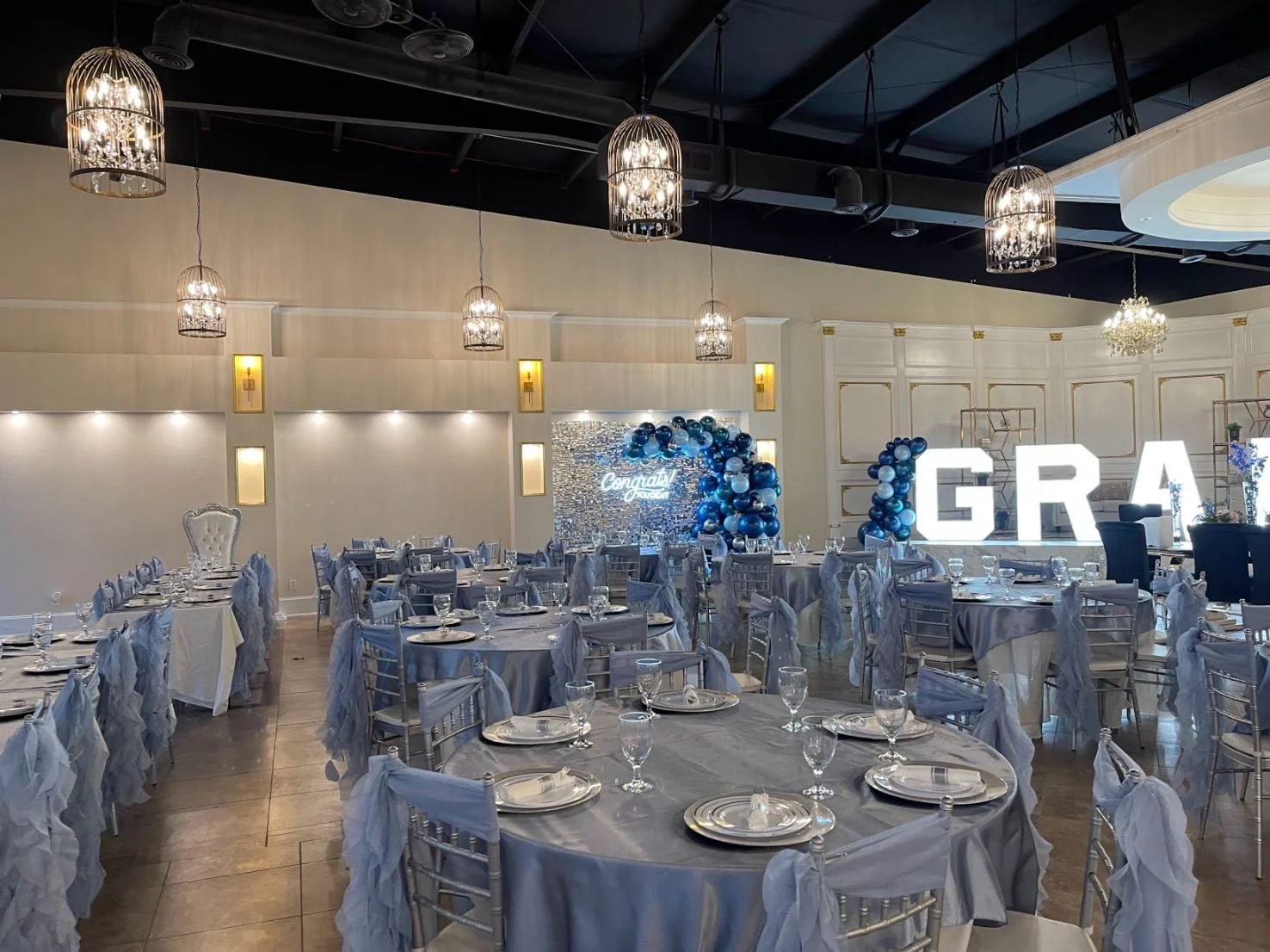  banquet hall decorated for a celebration with round tables covered in gray tablecloths, chairs with gray fabric sashes, glasses and plates set on each table, large illuminated 