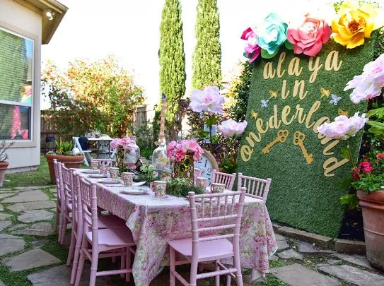 An outdoor dining setup with a long pink tablecloth-covered table and matching pink chairs arranged on a stone patio. The table is decorated with glass jars filled with pink flowers, and the background features a green wall with colorful paper flower