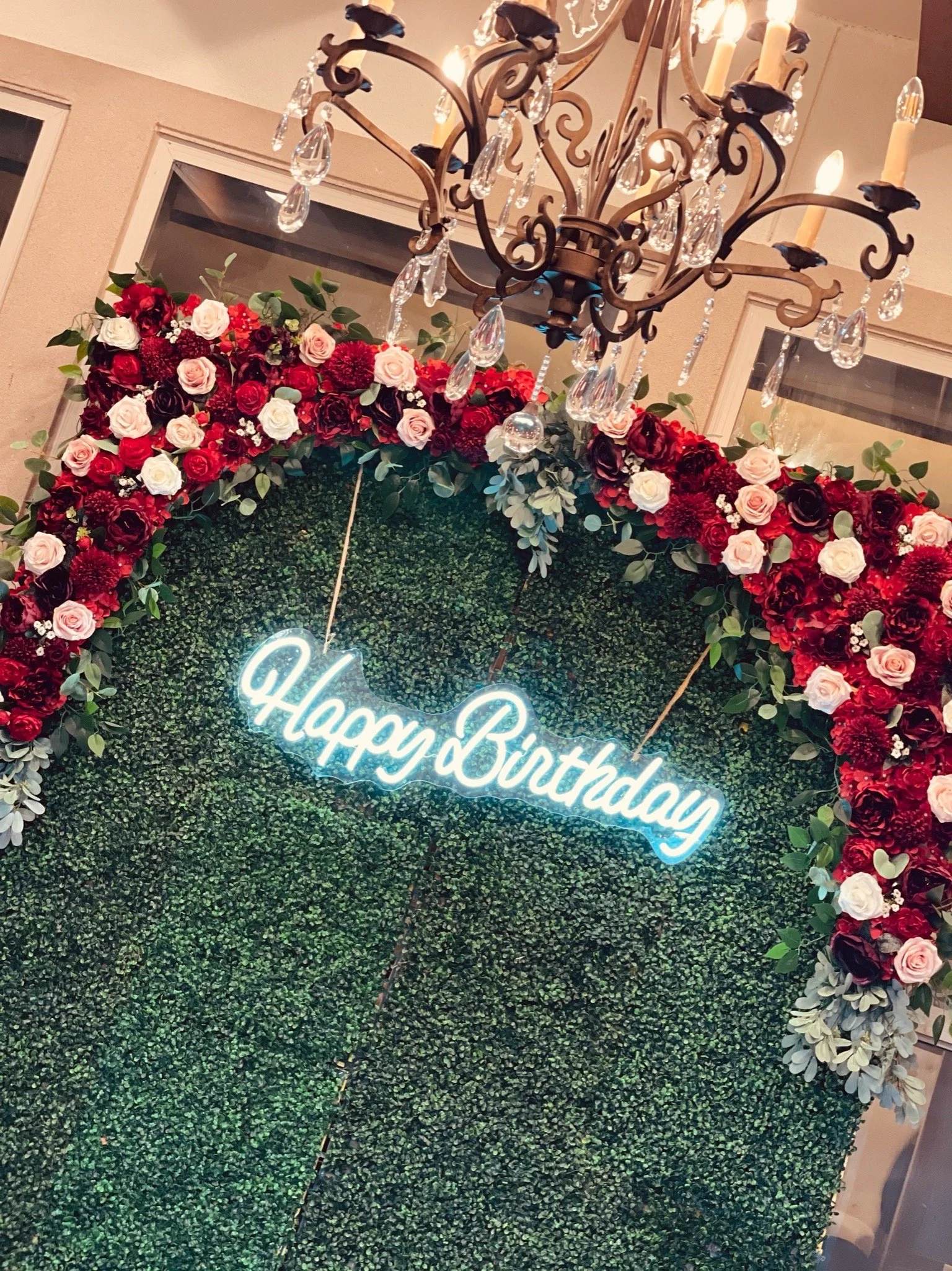 A decorative backdrop with a dense green leafy wall, surrounded by an arrangement of red, pink, and white flowers, with a neon sign that reads 'Happy Birthday' hanging in the center. A chandelier with ornate metalwork and dangling crystals is above.