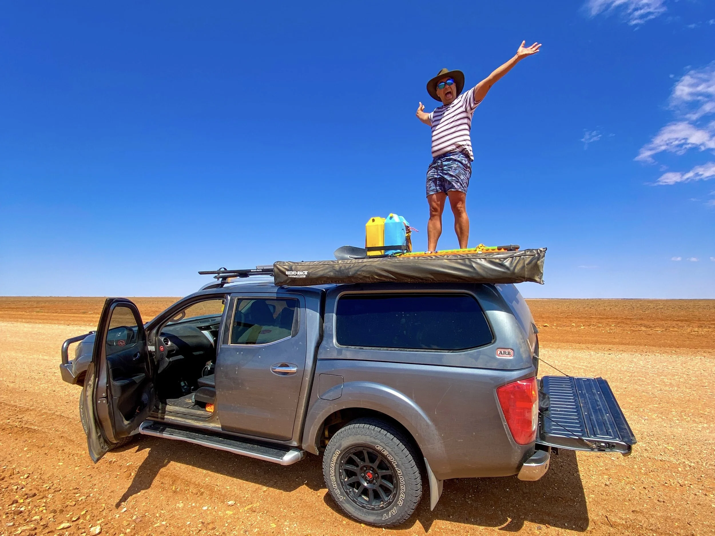Person standing on top of a ute with arms outstretched on a sunny day surrounded by red dirt plains