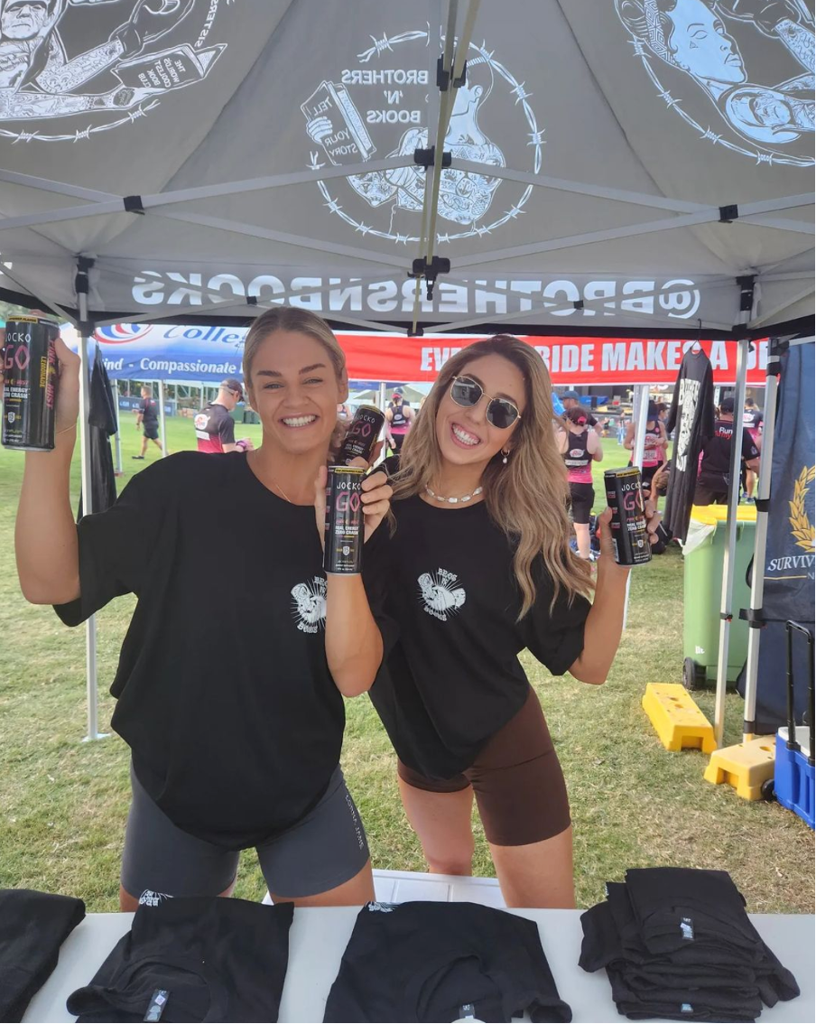 Two women holding energy drink cans at a booth under a canopy with branded shirts and signage.