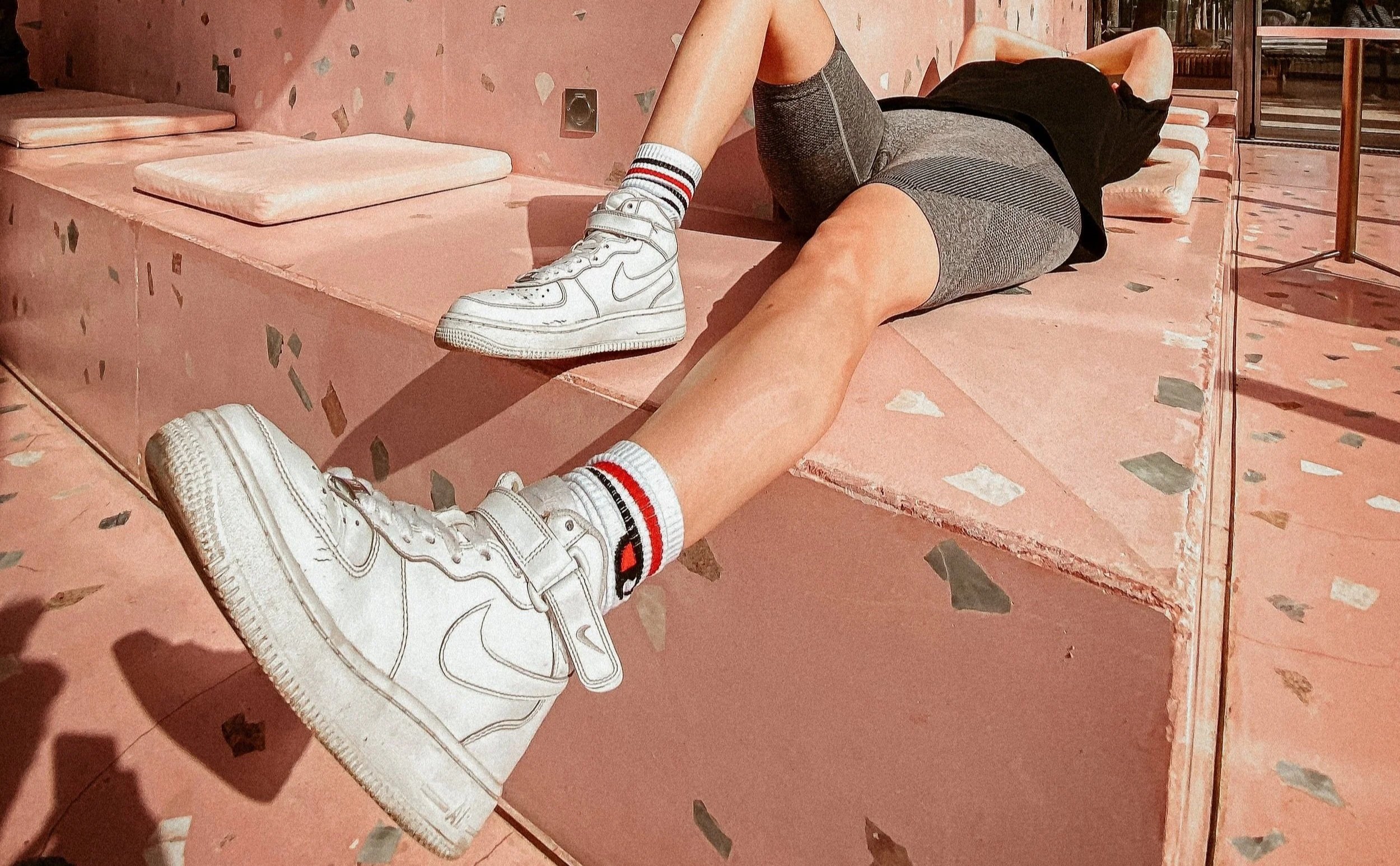 Persoon met sportkleding en witte sneakers ligt op een roze bank met gekleurde tegels op de vloer.