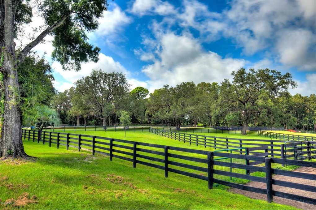 A lush green pasture with black wooden fencing, large oak trees, and a partly cloudy sky.