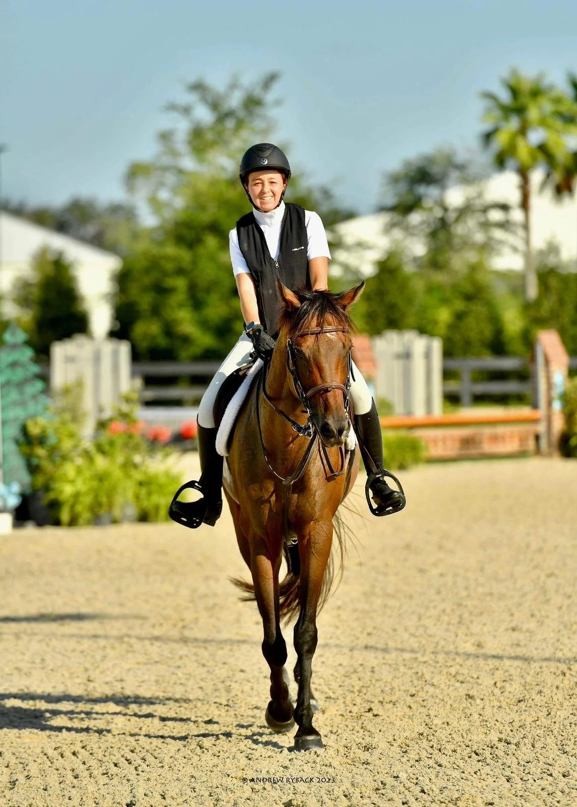 A woman riding a brown horse in an outdoor riding arena. The woman is smiling, wearing a black helmet and riding gear, and the background features green trees and a fence.