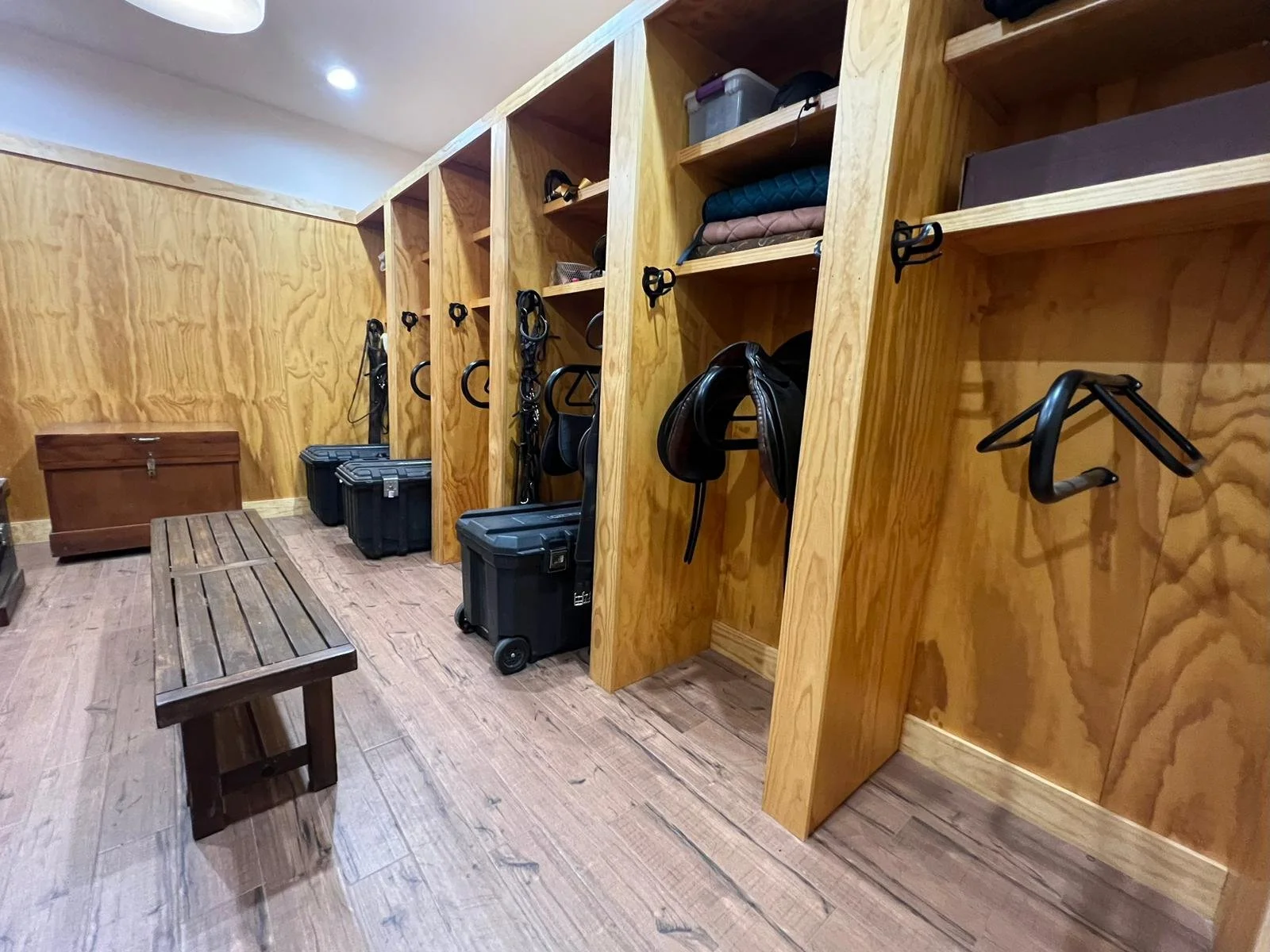 A wooden tack room with open compartments containing horse tack and gear, a bench, and a wooden chest in the background.